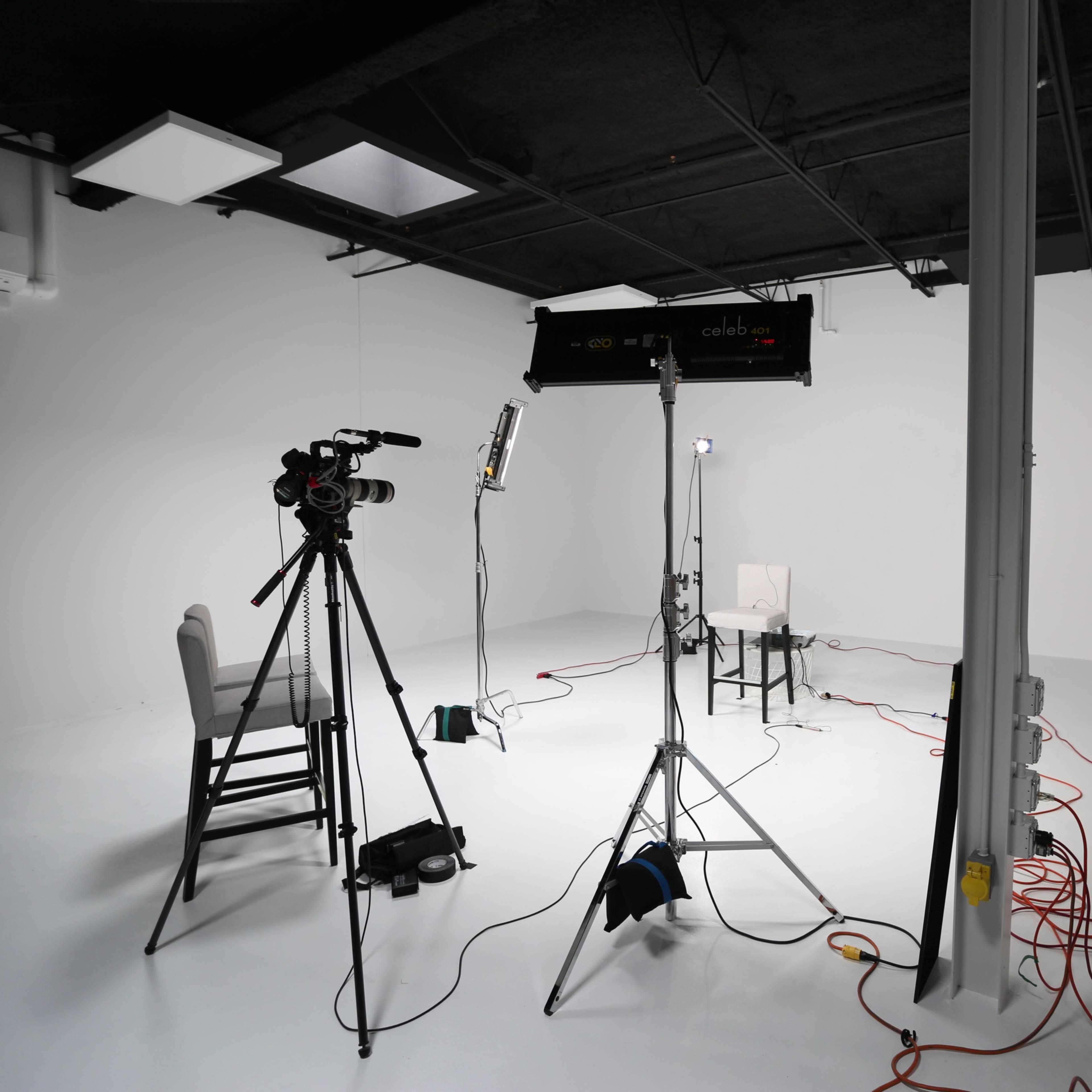 A video production setup features a camera, lighting equipment, and a single chair in an empty white studio.