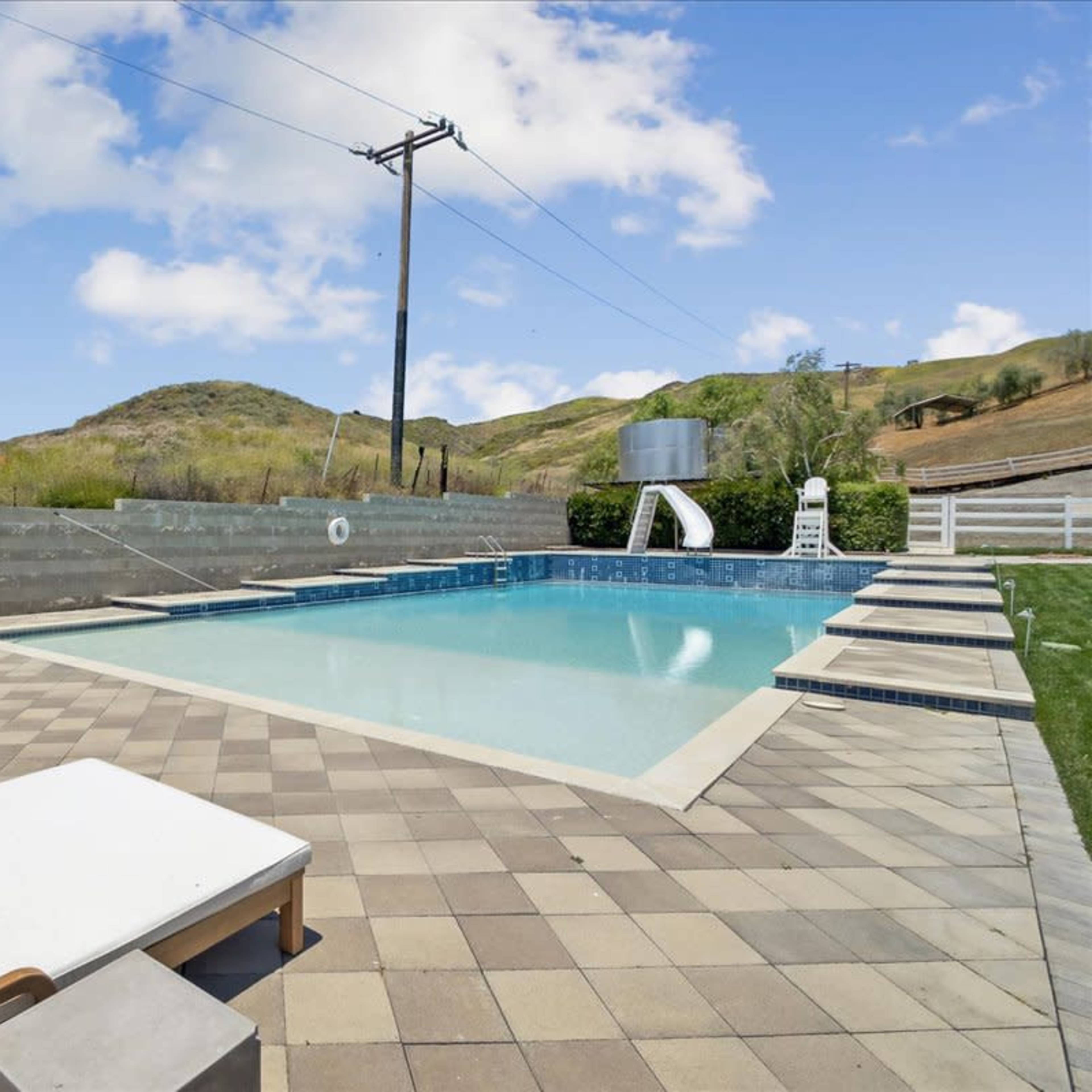 A swimming pool with tiled decking is surrounded by lounge chairs and overlooks rolling hills under a clear sky.