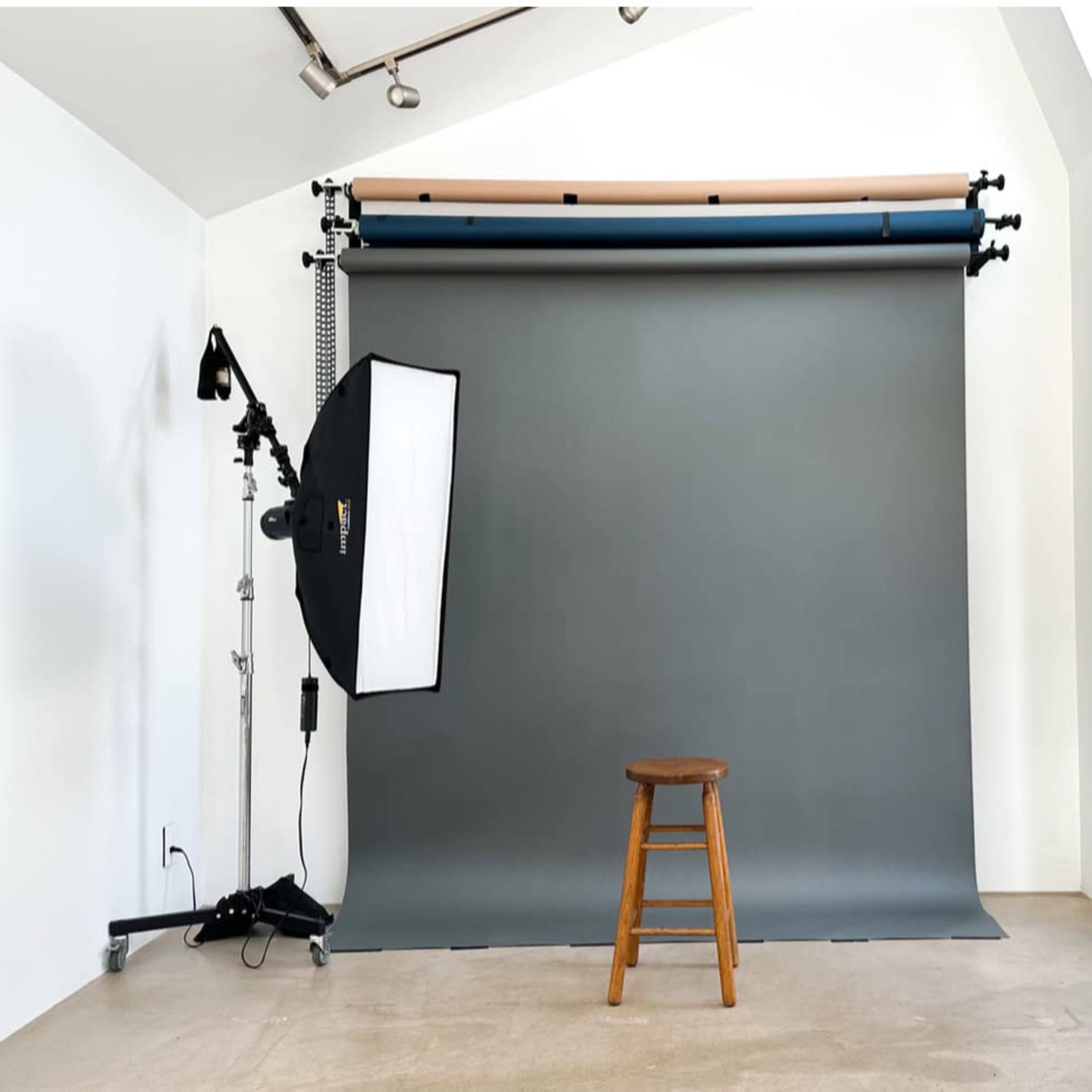 A photography studio features a gray backdrop, a wooden stool, and a lighting setup on the left side.