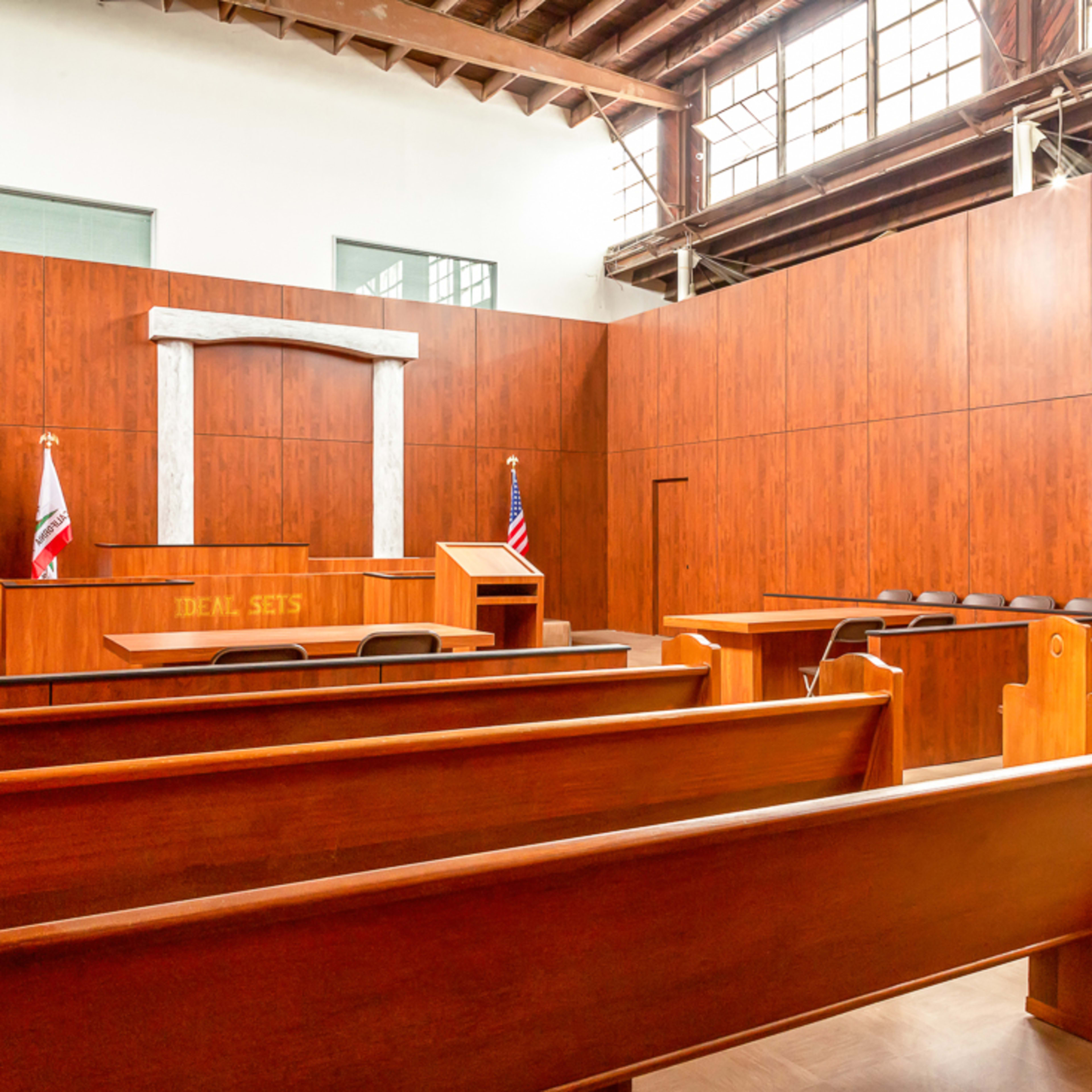 A wooden-paneled courtroom with empty benches, a judge's bench, and flags at the front.