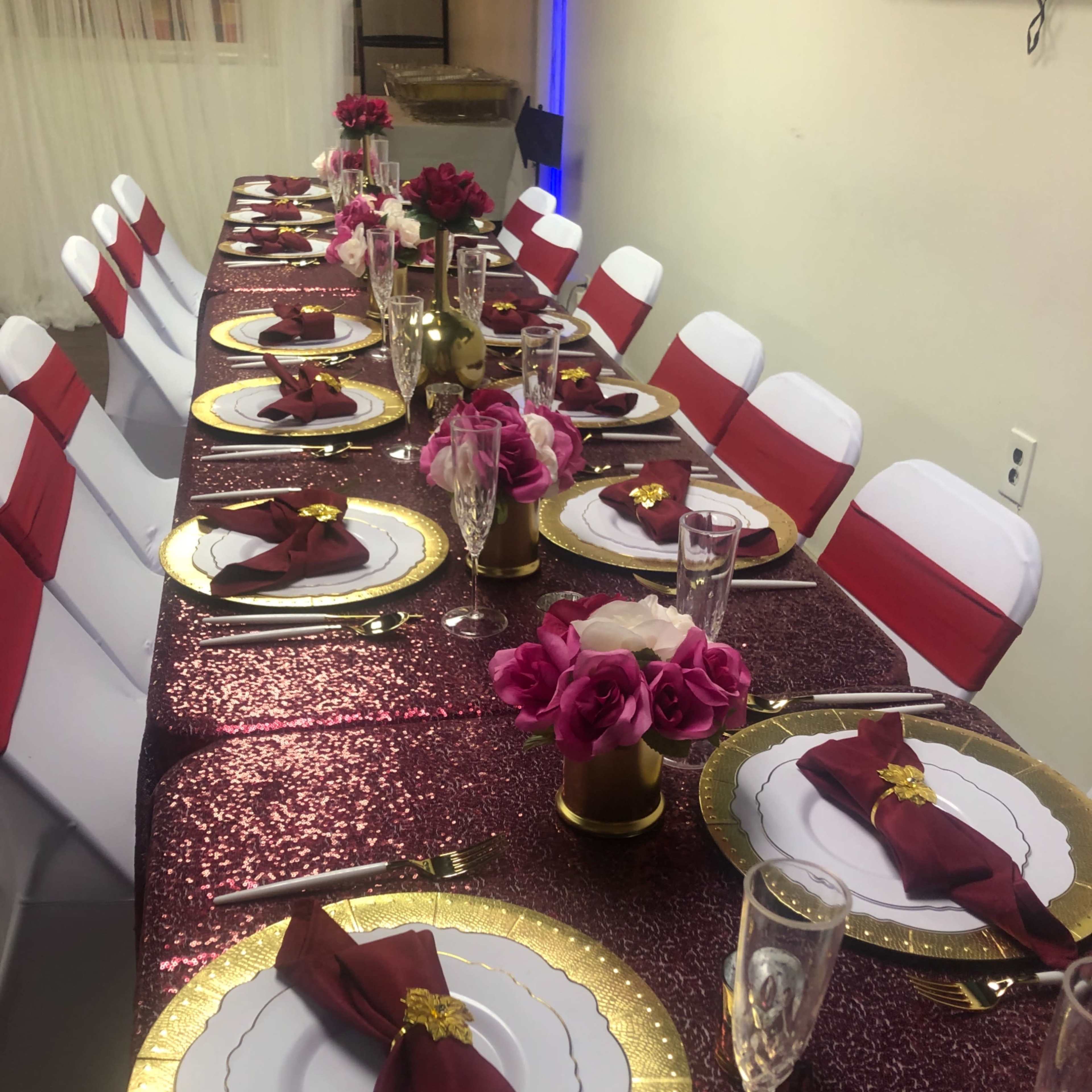 A long dining table is set with gold-rimmed plates, burgundy napkins, pink floral centerpieces, and shimmering tablecloths, arranged for a formal event.