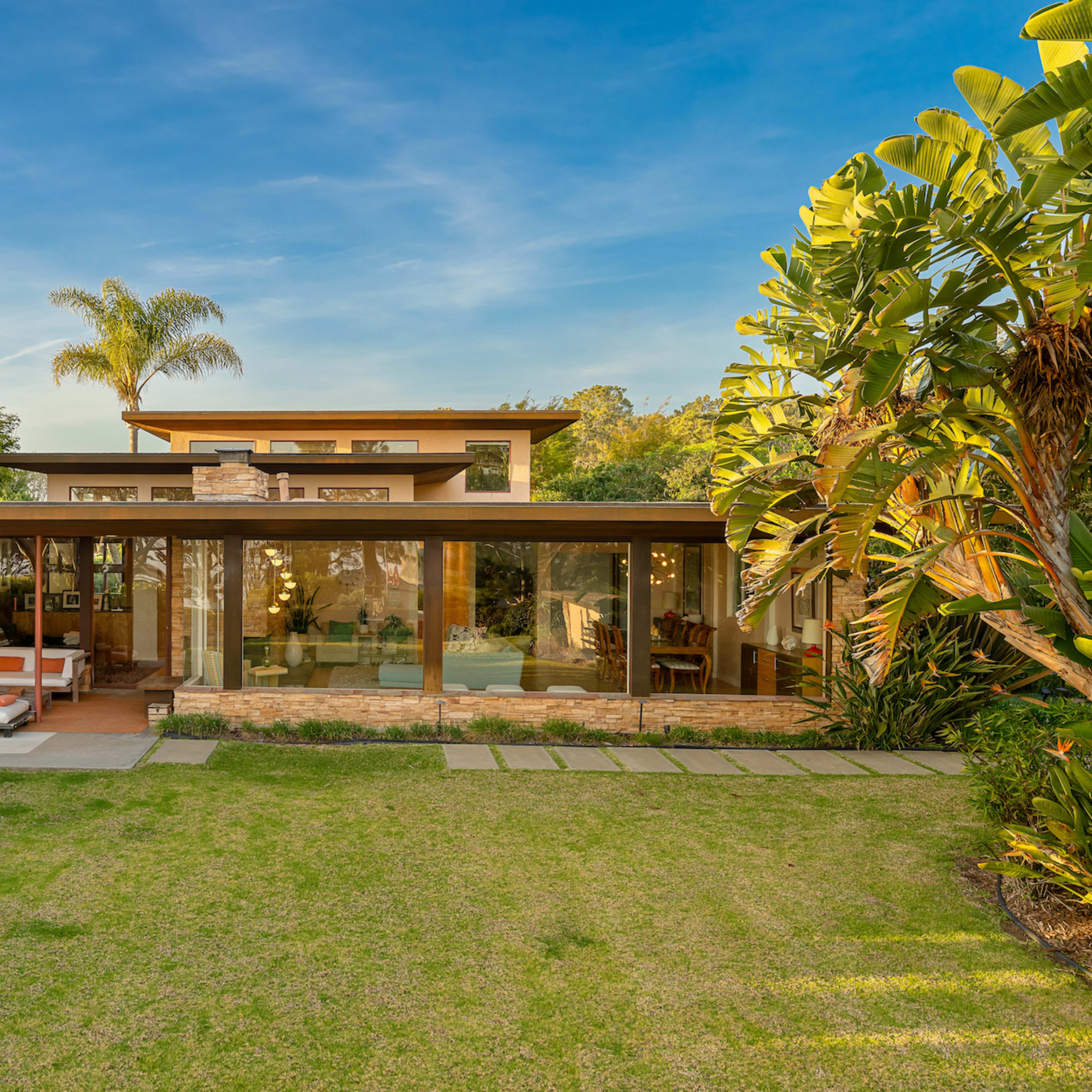 A modern house with large glass windows and a covered patio is surrounded by landscaped greenery.