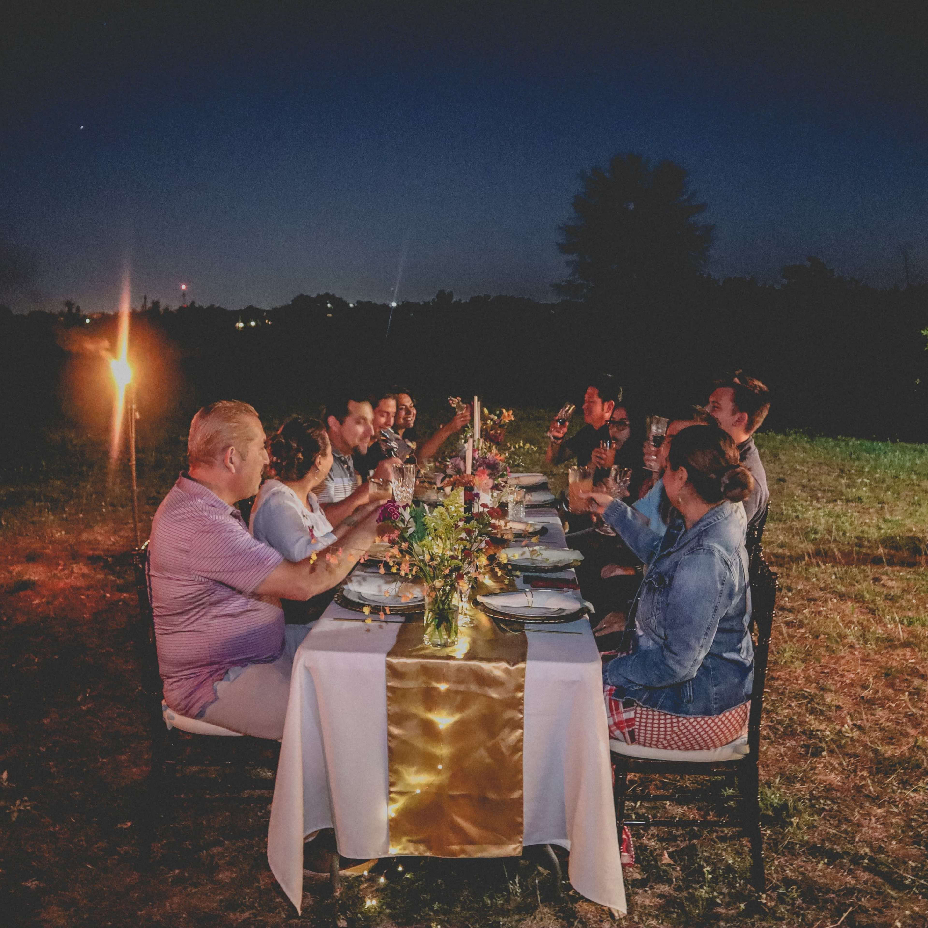 A group of people sits around a long table set for dinner in an outdoor setting at twilight, with tiki torches providing light and a floral centerpiece on the table.