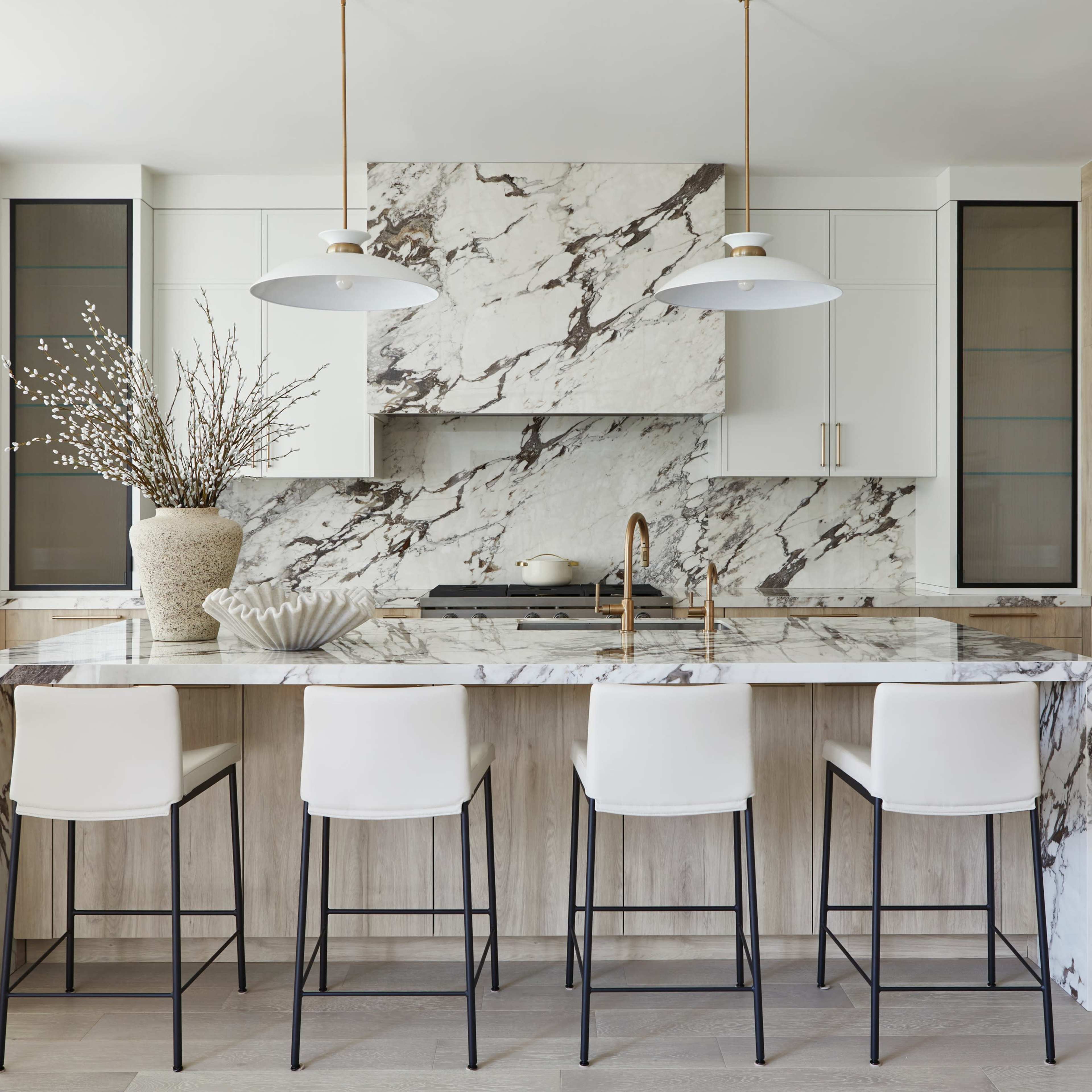A modern kitchen features a large marble island with bar stools, white cabinetry, and glass-fronted storage, accented by decorative branches in a vase.