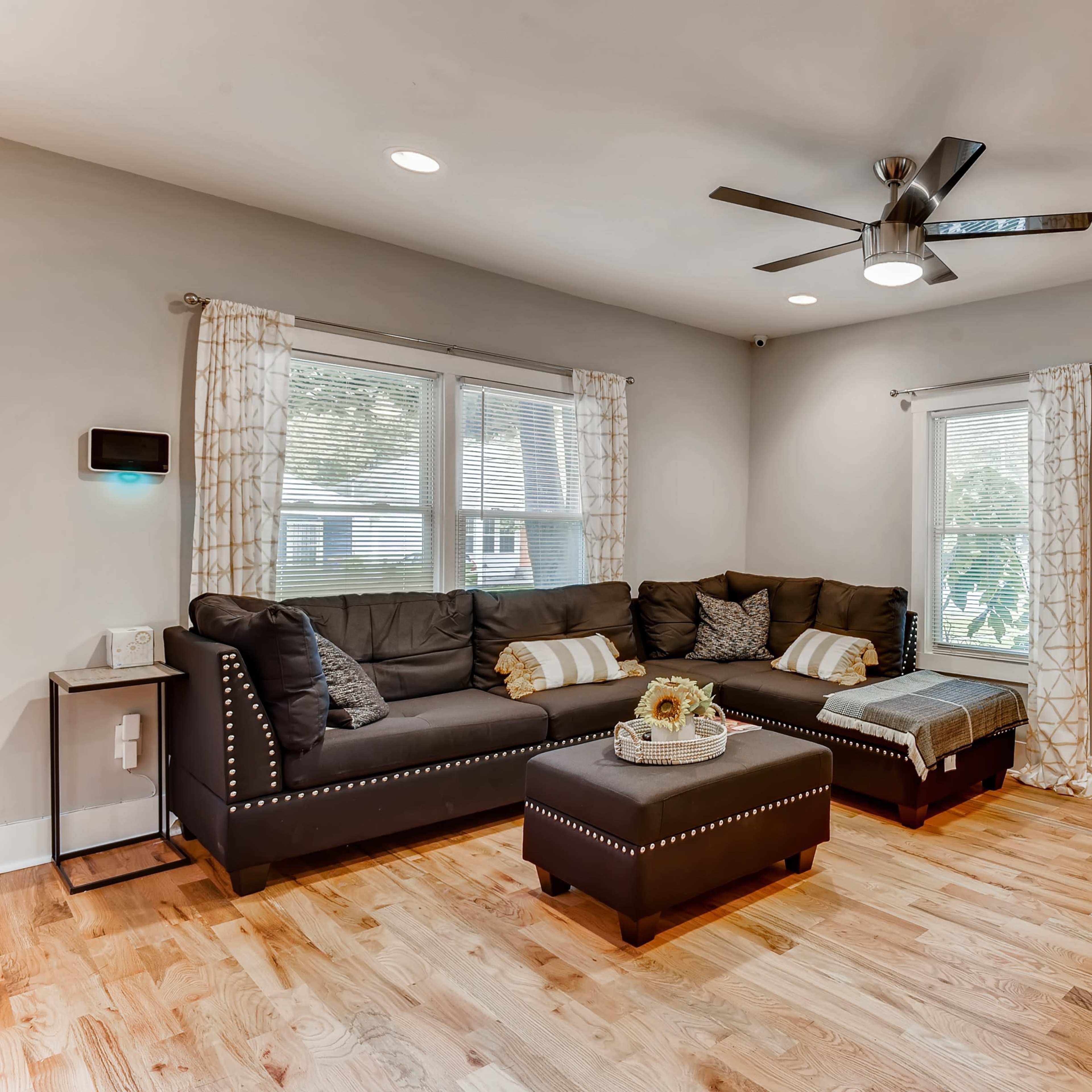 A modern living room features a sectional sofa, a coffee table with a floral arrangement, and a ceiling fan, with natural light coming through the windows.
