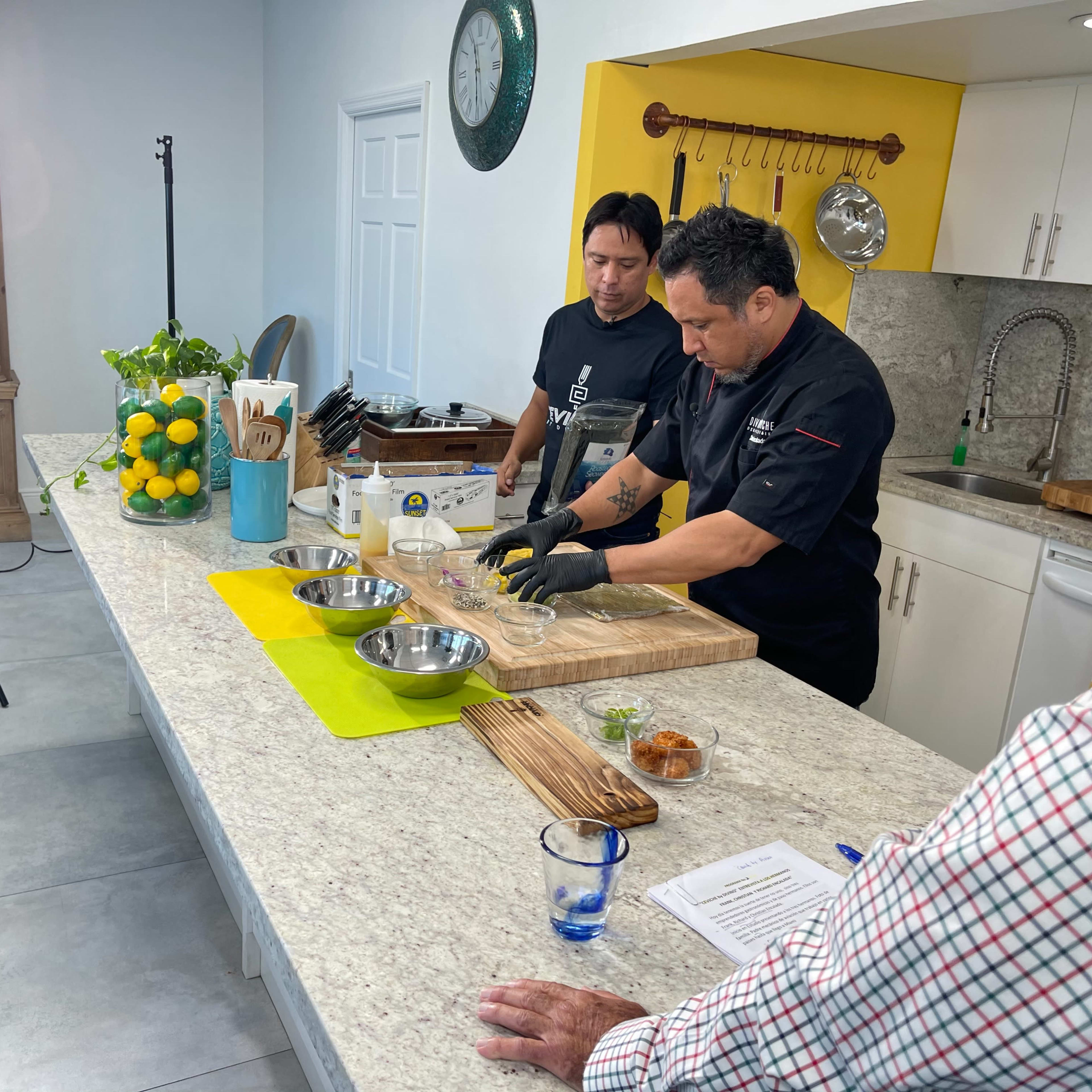 A chef prepares ingredients on a kitchen counter for a cooking demonstration while two observers watch and recording equipment is set up nearby.