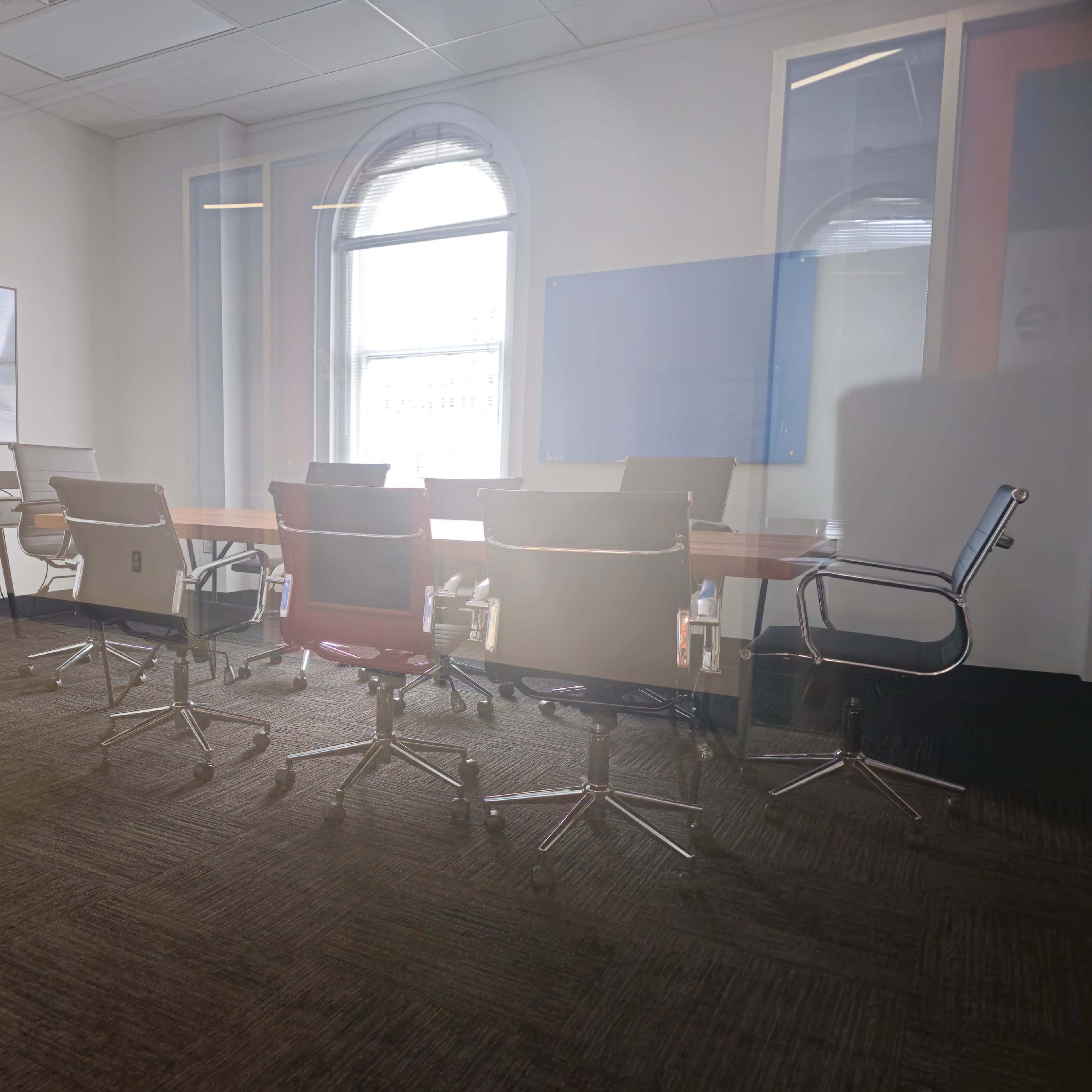 A modern conference room features a long wooden table surrounded by several rolling chairs and a large window providing natural light.