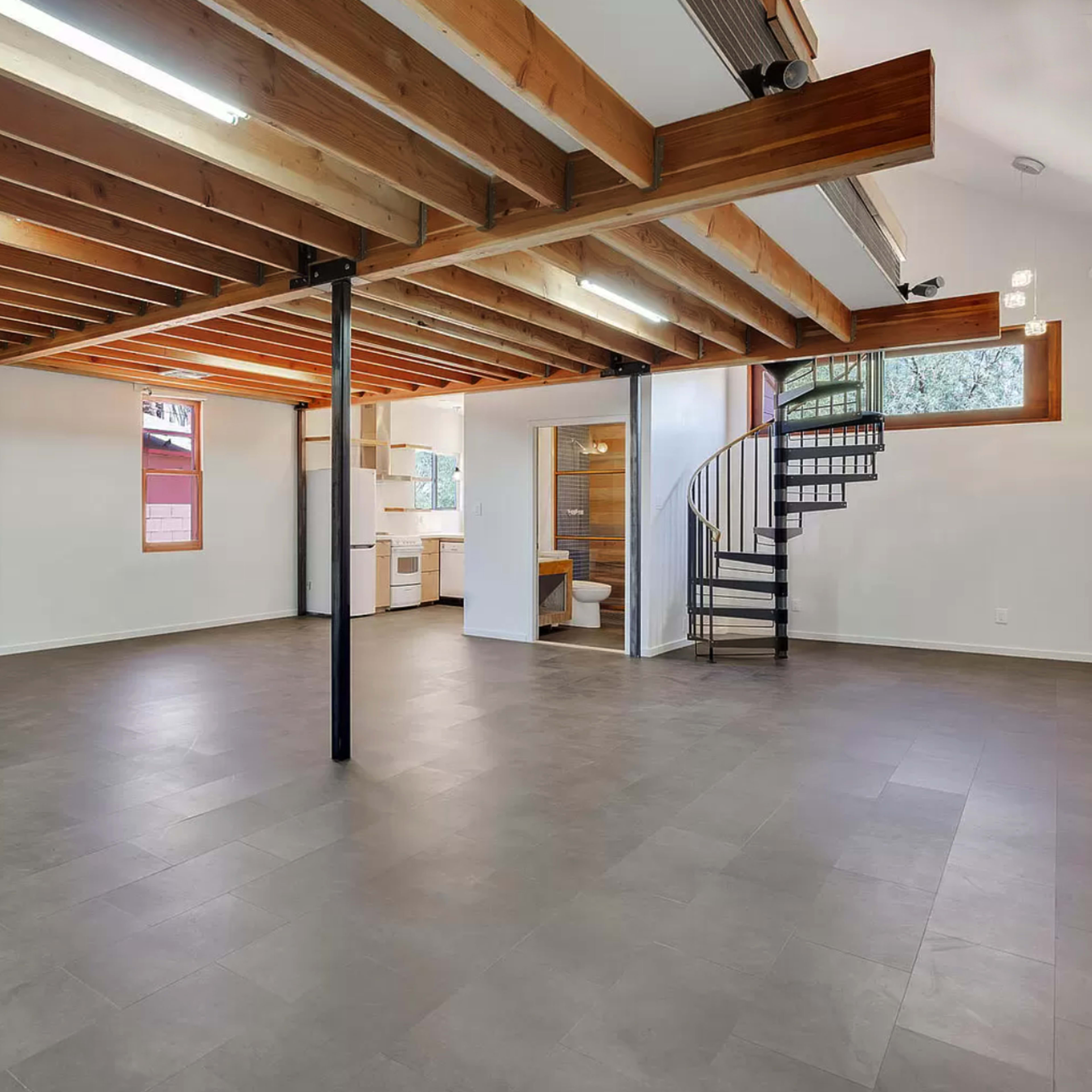 A spacious interior with exposed wooden beams, a spiral staircase, and a partially visible kitchen area in a modern basement layout.