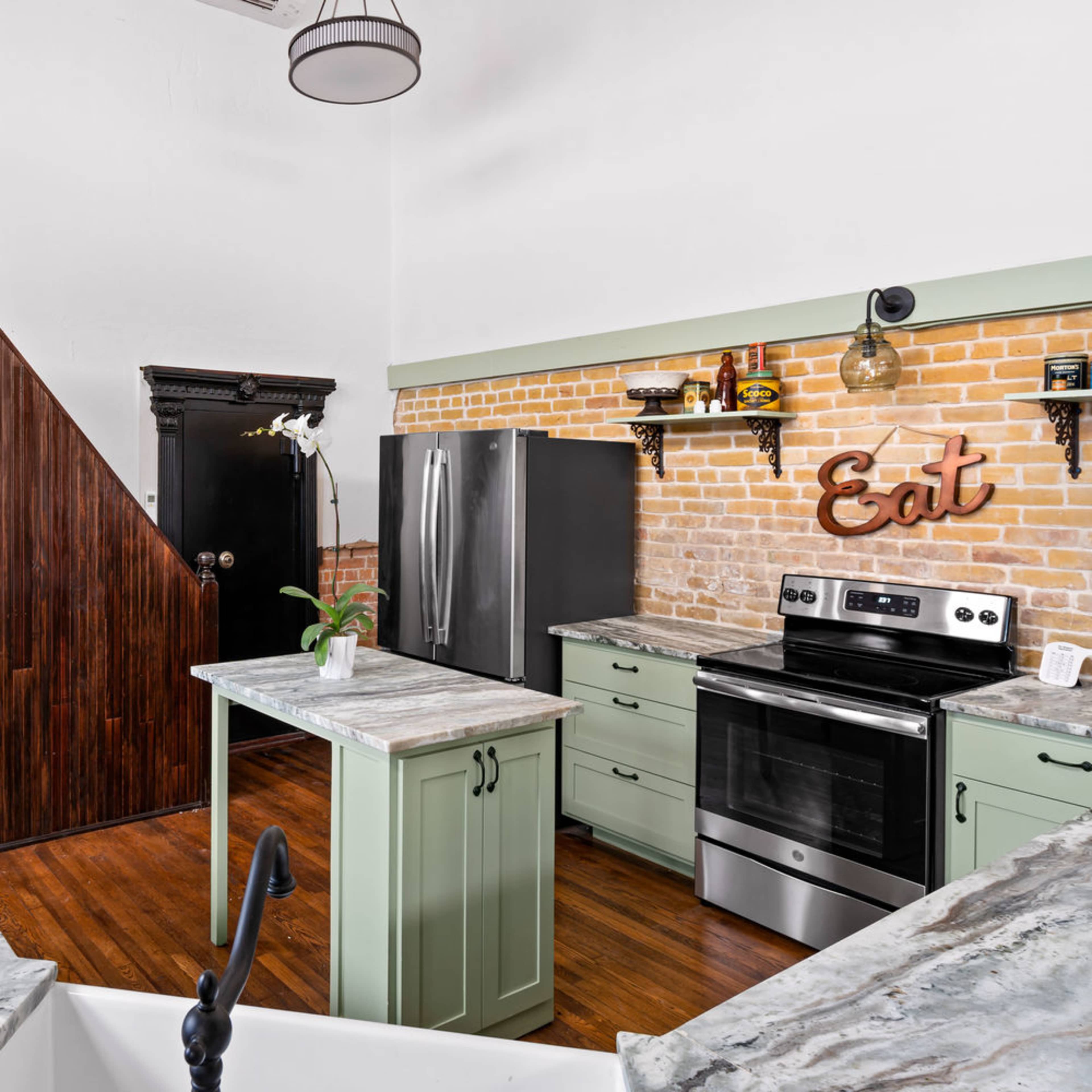 A modern kitchen with a central island, stainless steel appliances, and a brick wall, adjacent to a staircase leading to an upper level.