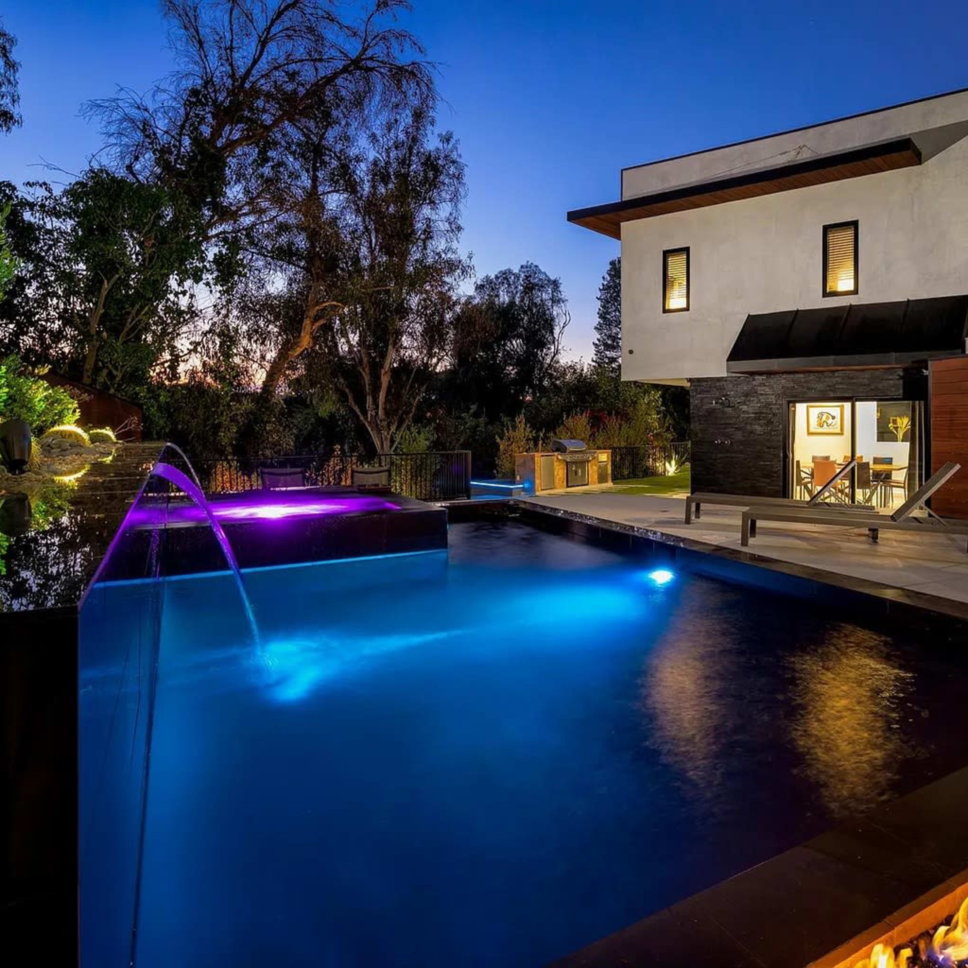 A modern two-story house is illuminated at dusk, showcasing a pool with colored lighting and a patio area.
