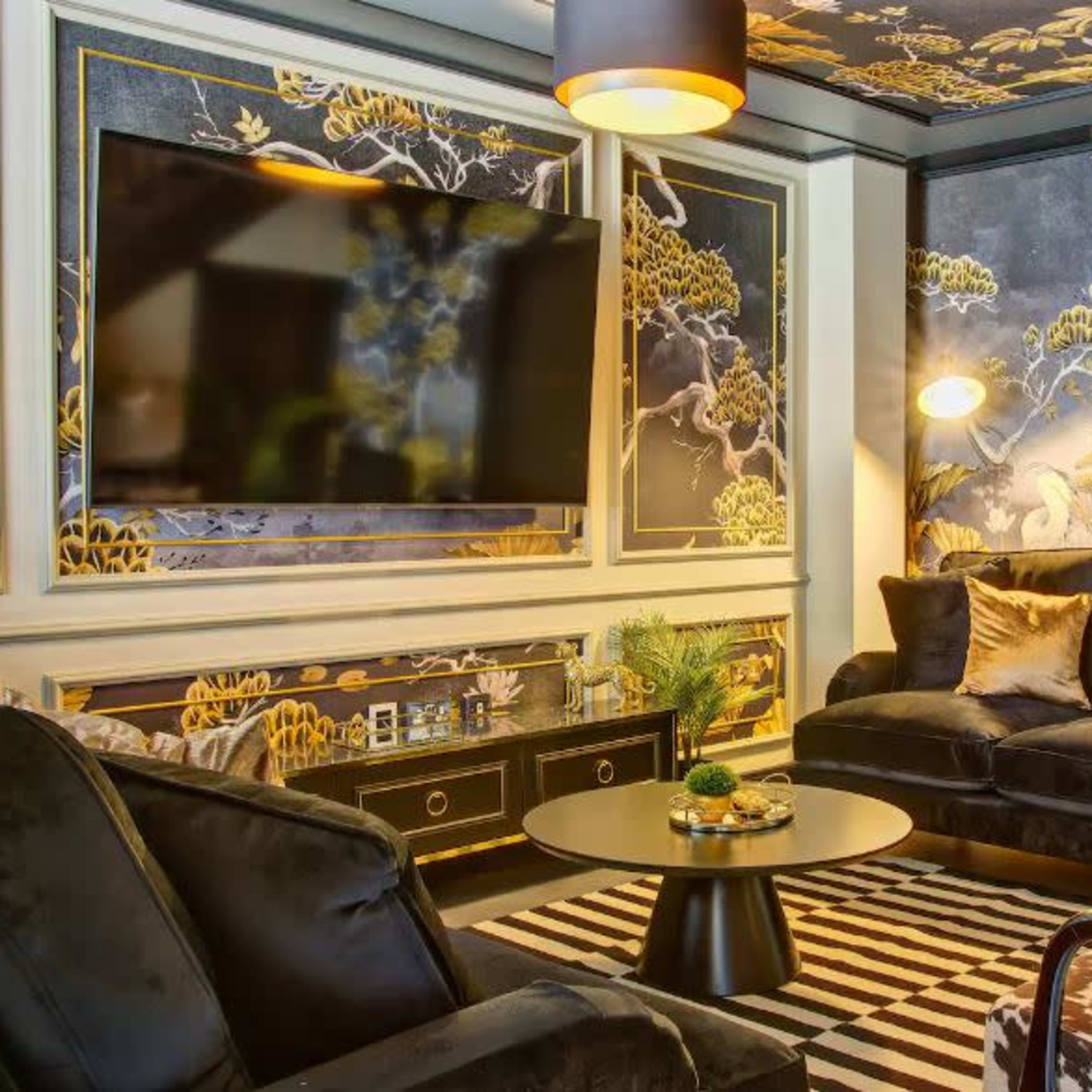 A modern living room with a large flat-screen TV, two black sofas, and a coffee table, all set against a wall adorned with a decorative mural and patterned wallpaper.