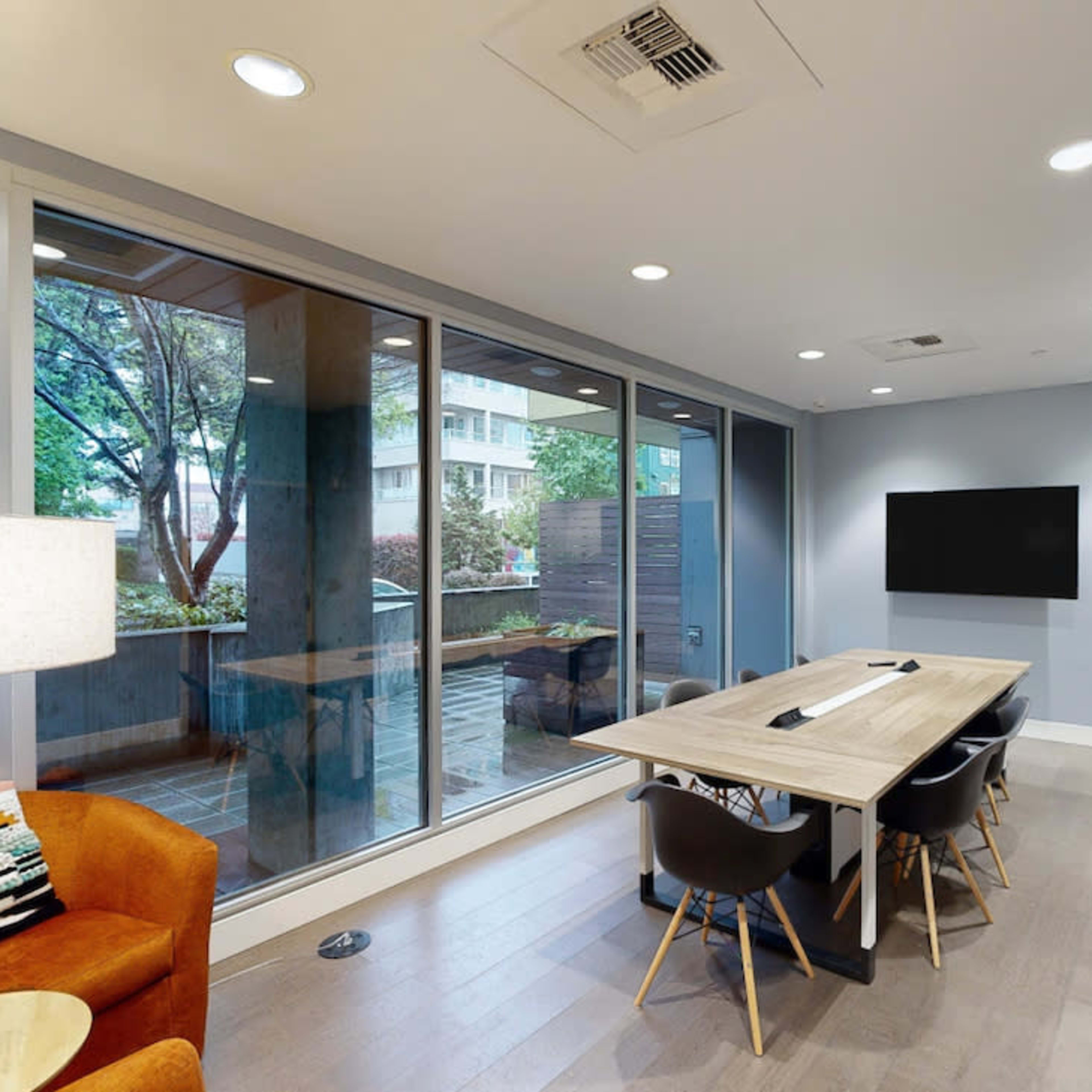 A modern conference room with a large wooden table, black chairs, and a wall-mounted TV, featuring large windows that provide a view of the outside.