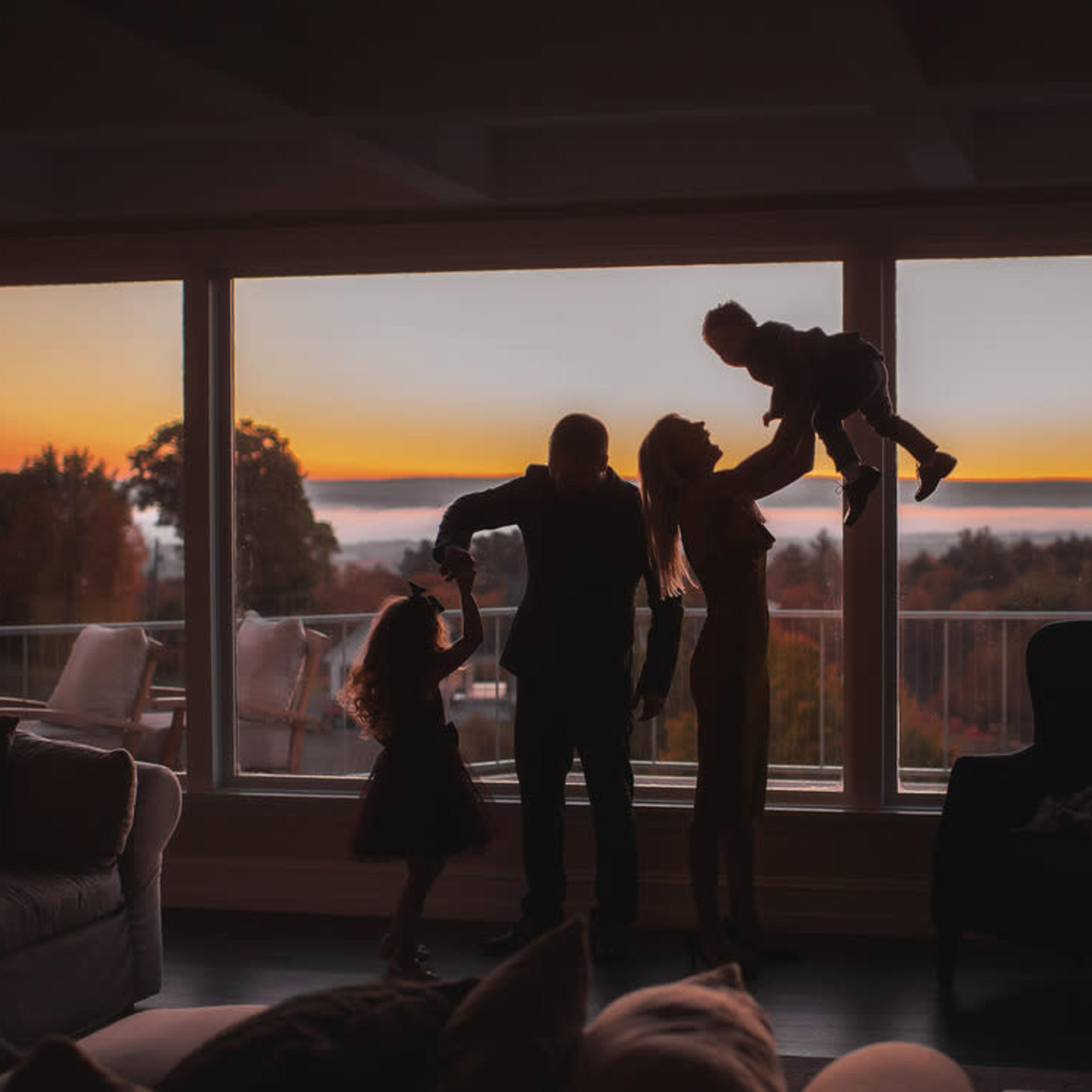 A family of four is silhouetted against a sunset seen through large windows.