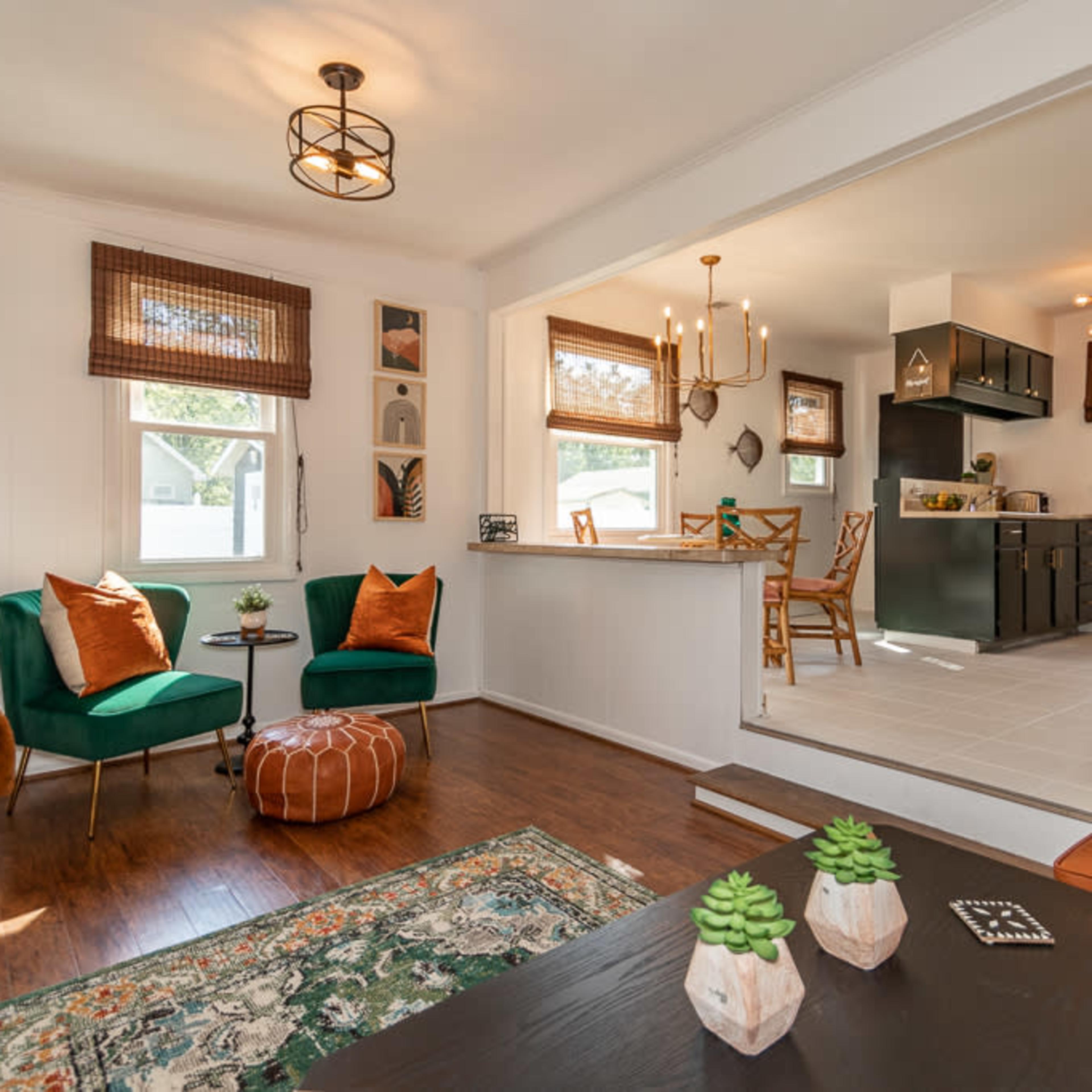 A cozy living area with two green chairs, a round ottoman, and a view into a kitchen featuring a dining area.