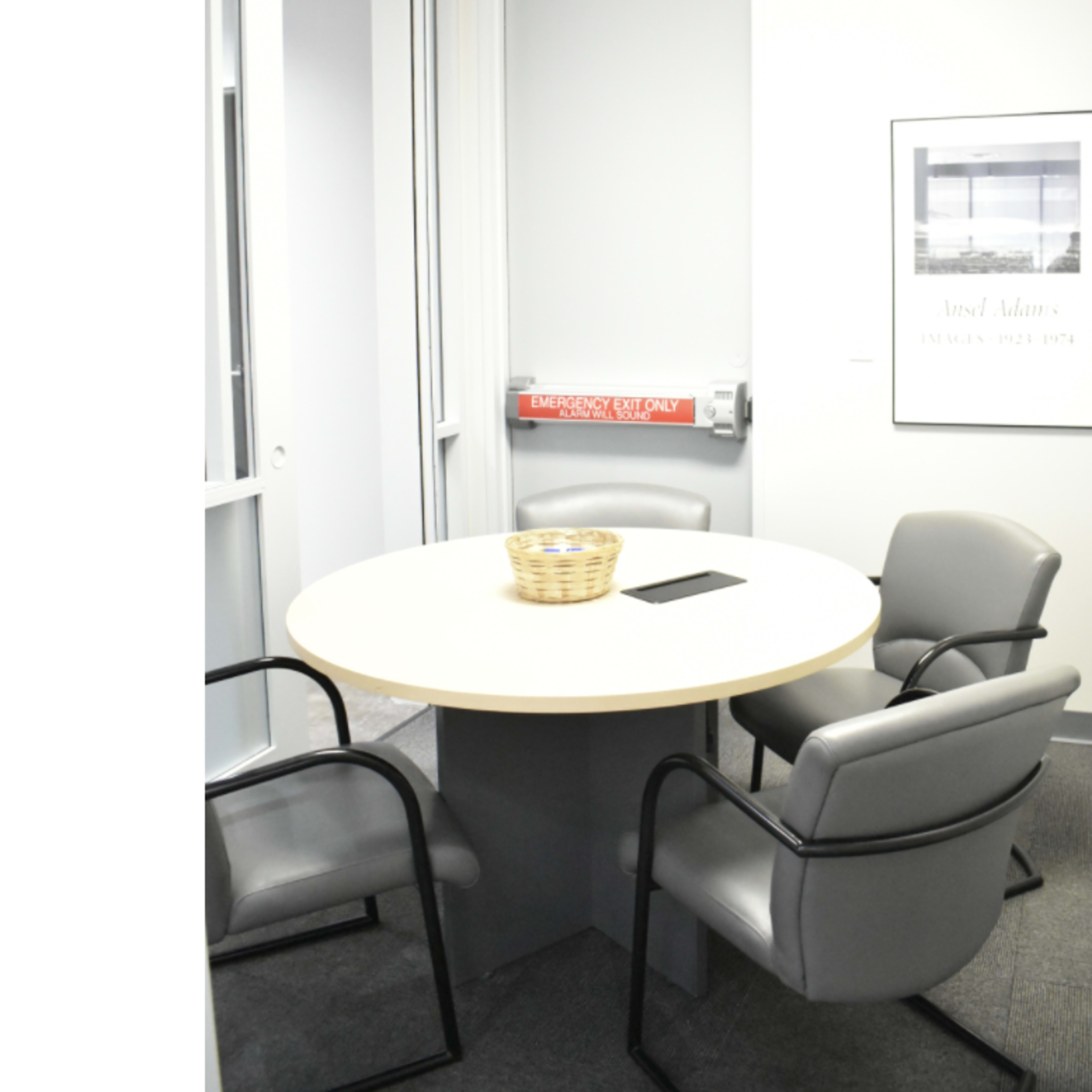 A round table with three gray chairs and a basket sits in a small office meeting room, featuring a framed picture on the wall and an emergency exit sign.
