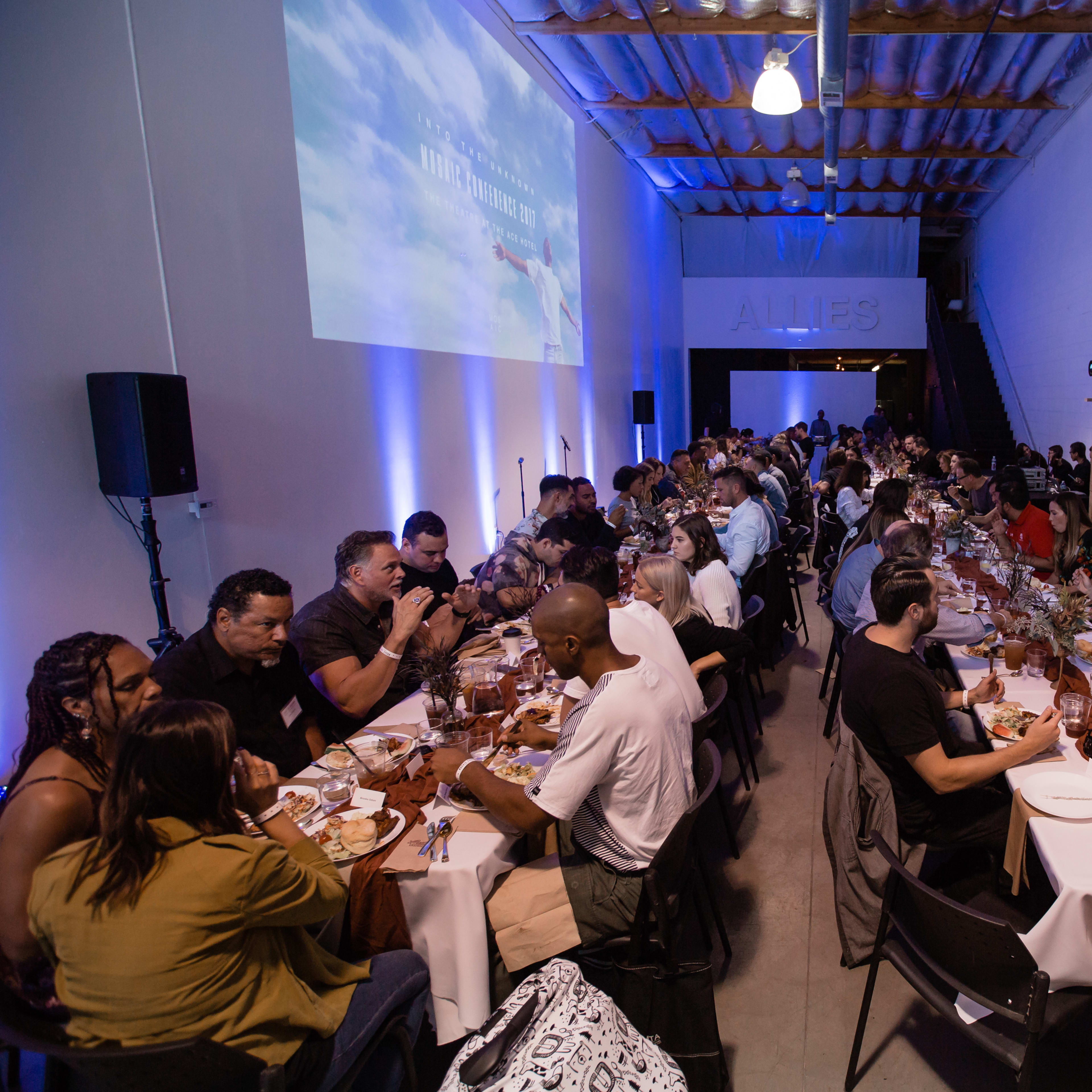 A large group of people is seated at long tables in a narrow event space, enjoying a meal together with a projector display in the background.