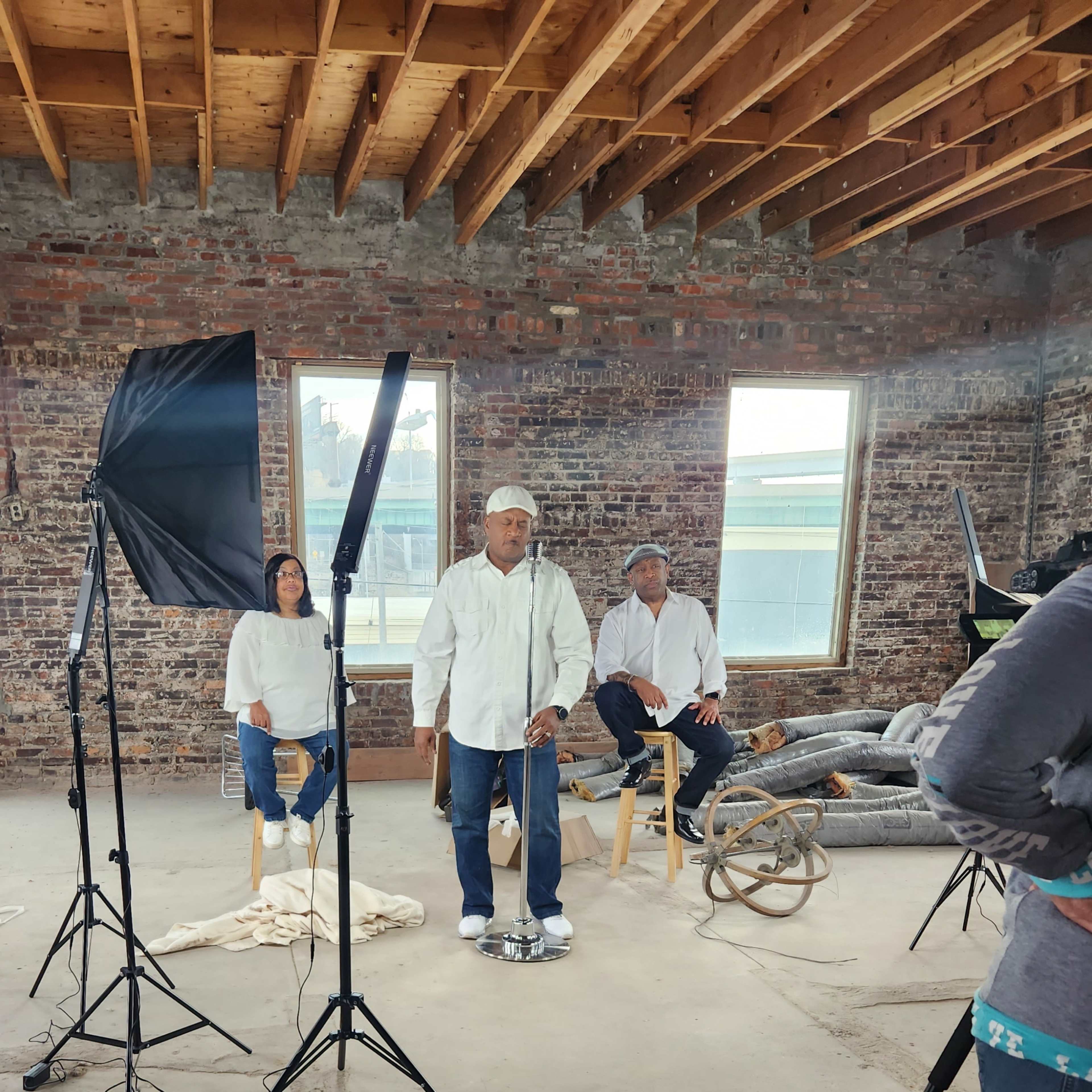 A group of three people poses for a photo shoot in a partially renovated industrial space with exposed brick walls and studio lighting setup.