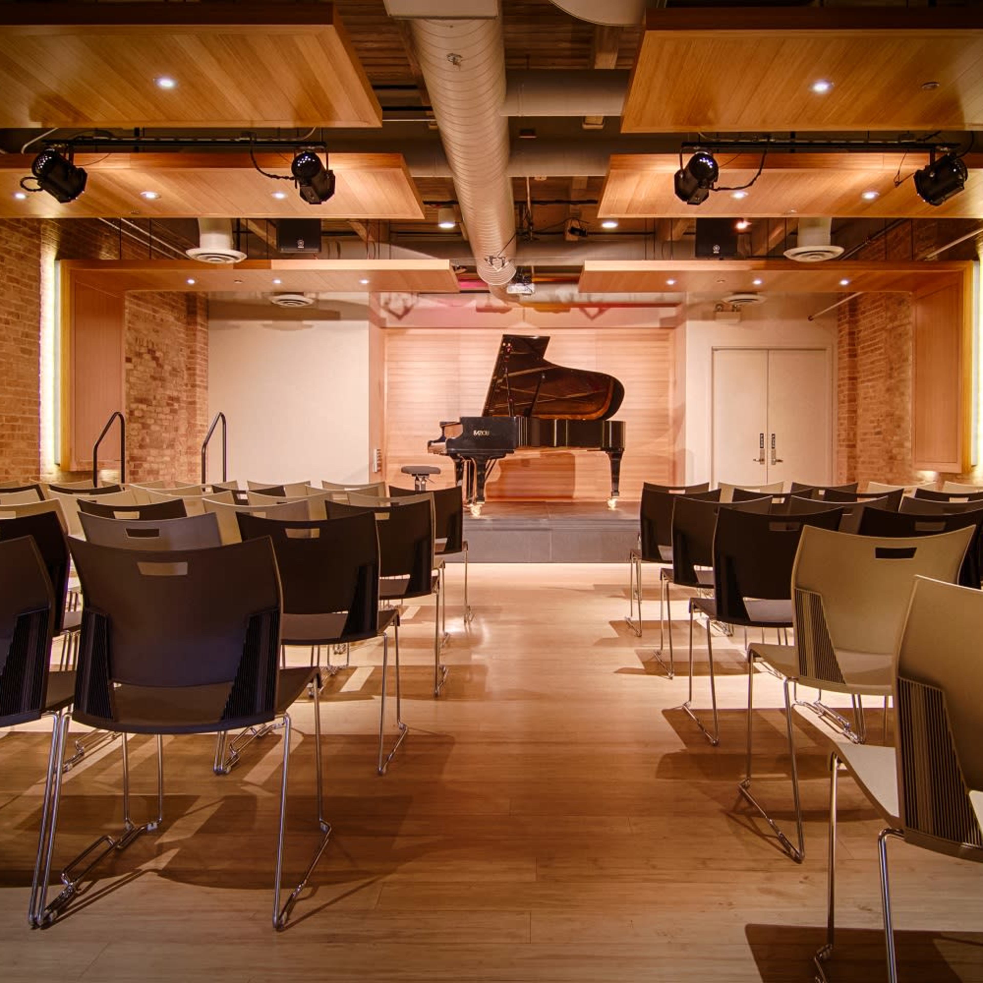 A concert hall with rows of chairs facing a grand piano on stage, surrounded by wooden beams and warm lighting.