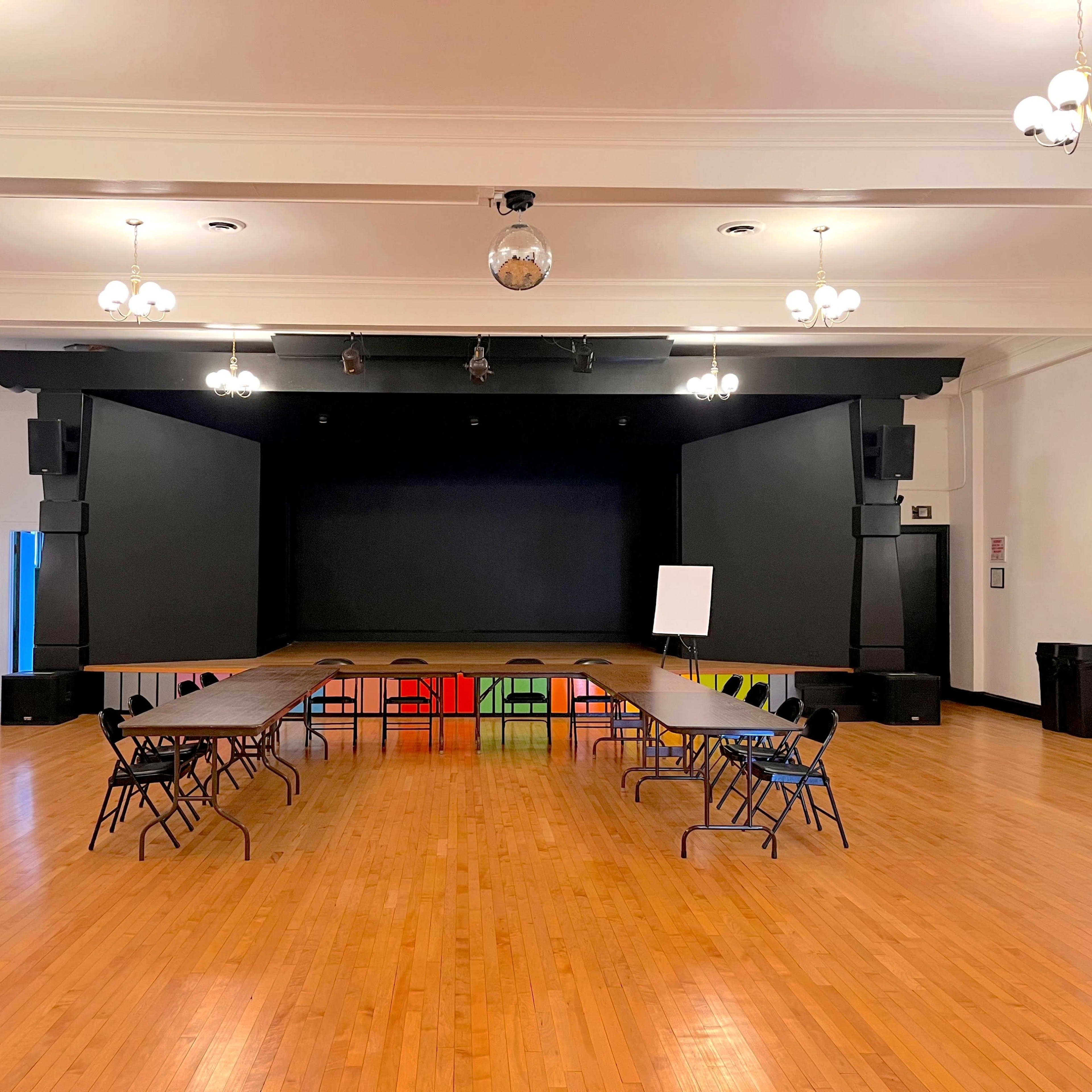 A spacious event hall with a stage at the front, featuring a row of tables arranged in a U-shape and chairs facing the stage.