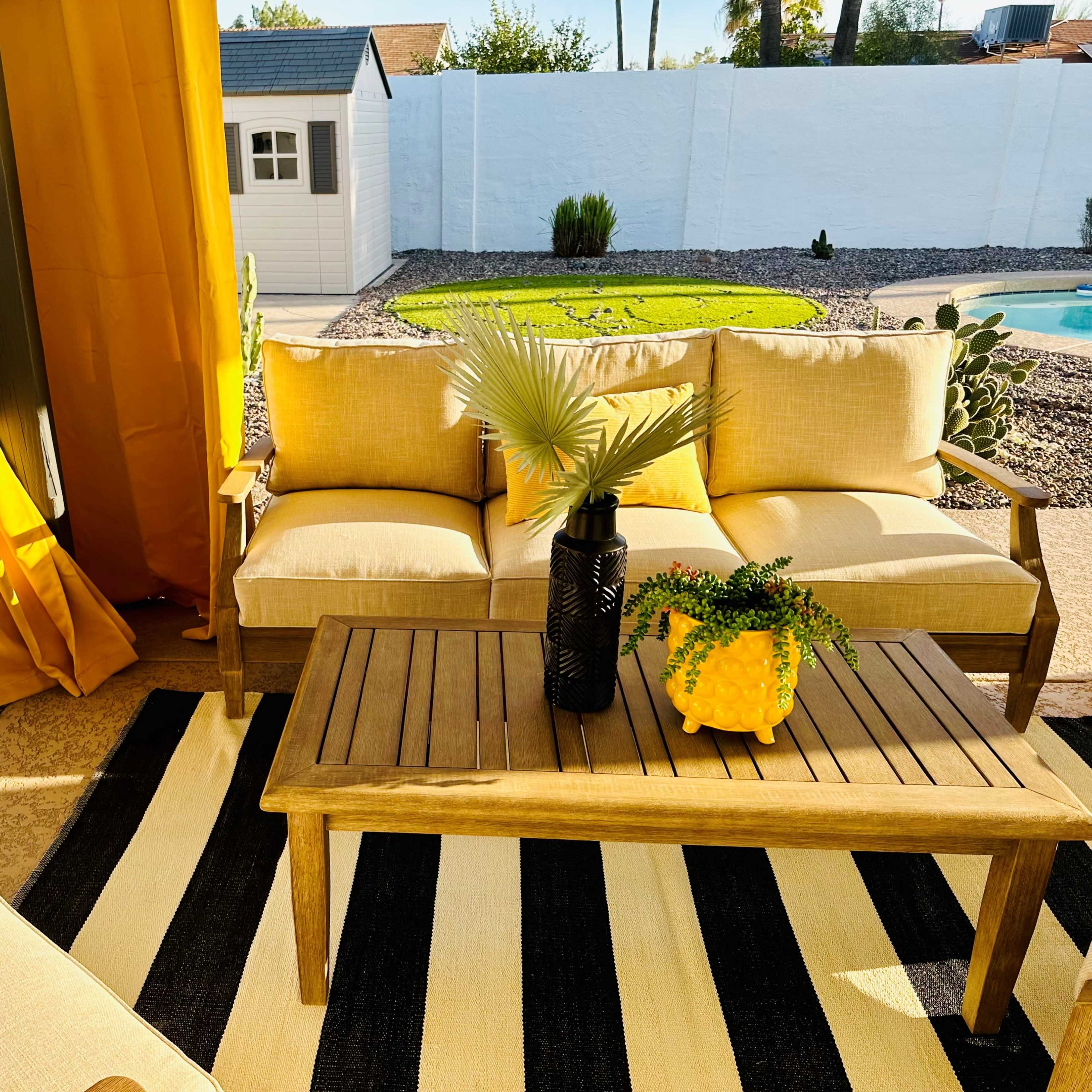A patio seating area features a wooden table and couch with yellow cushions, surrounded by a striped rug and yellow curtains, overlooking a landscaped yard.