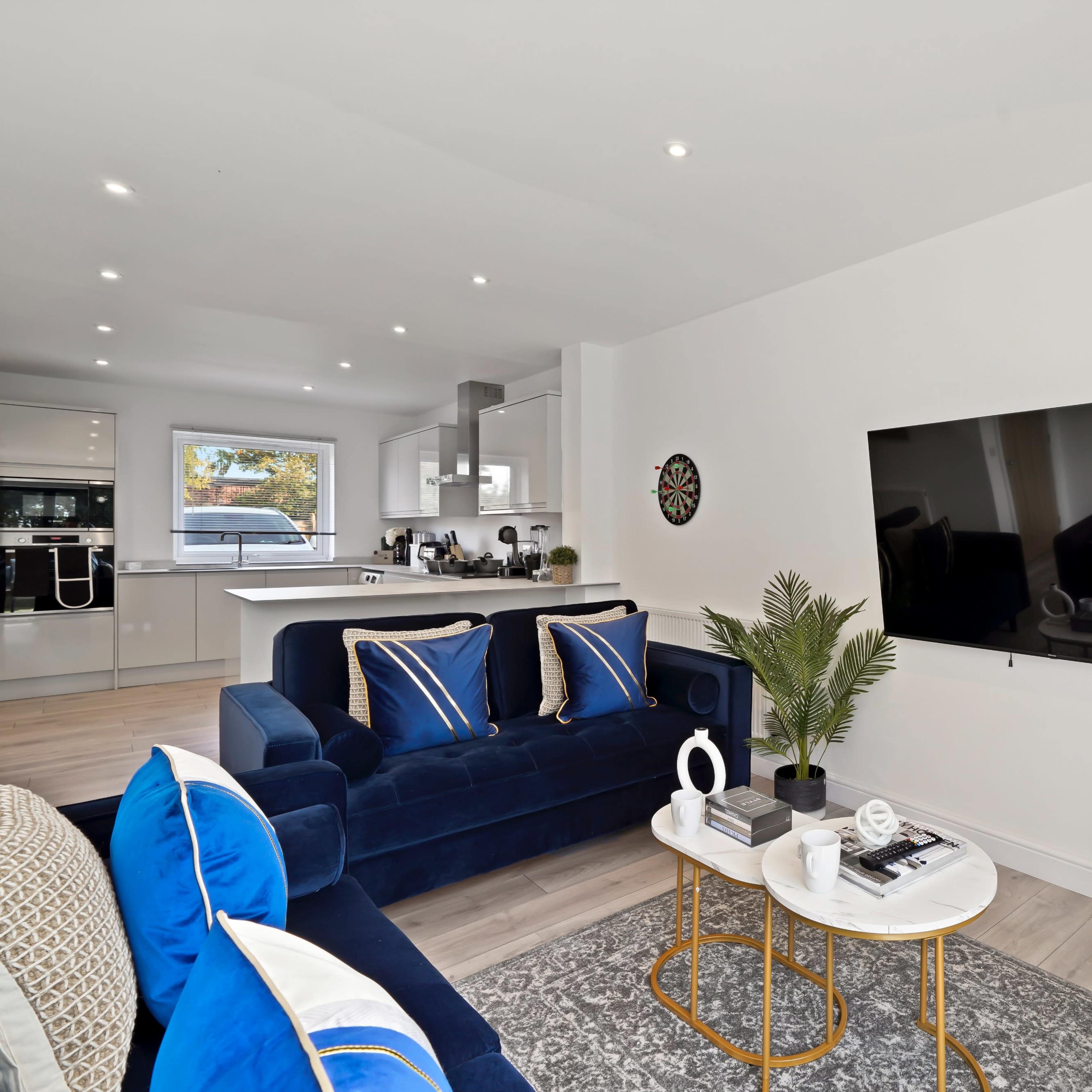A modern living room featuring a blue sofa, a wall-mounted television, and a small coffee table with decorative items, adjacent to a well-lit kitchen area.