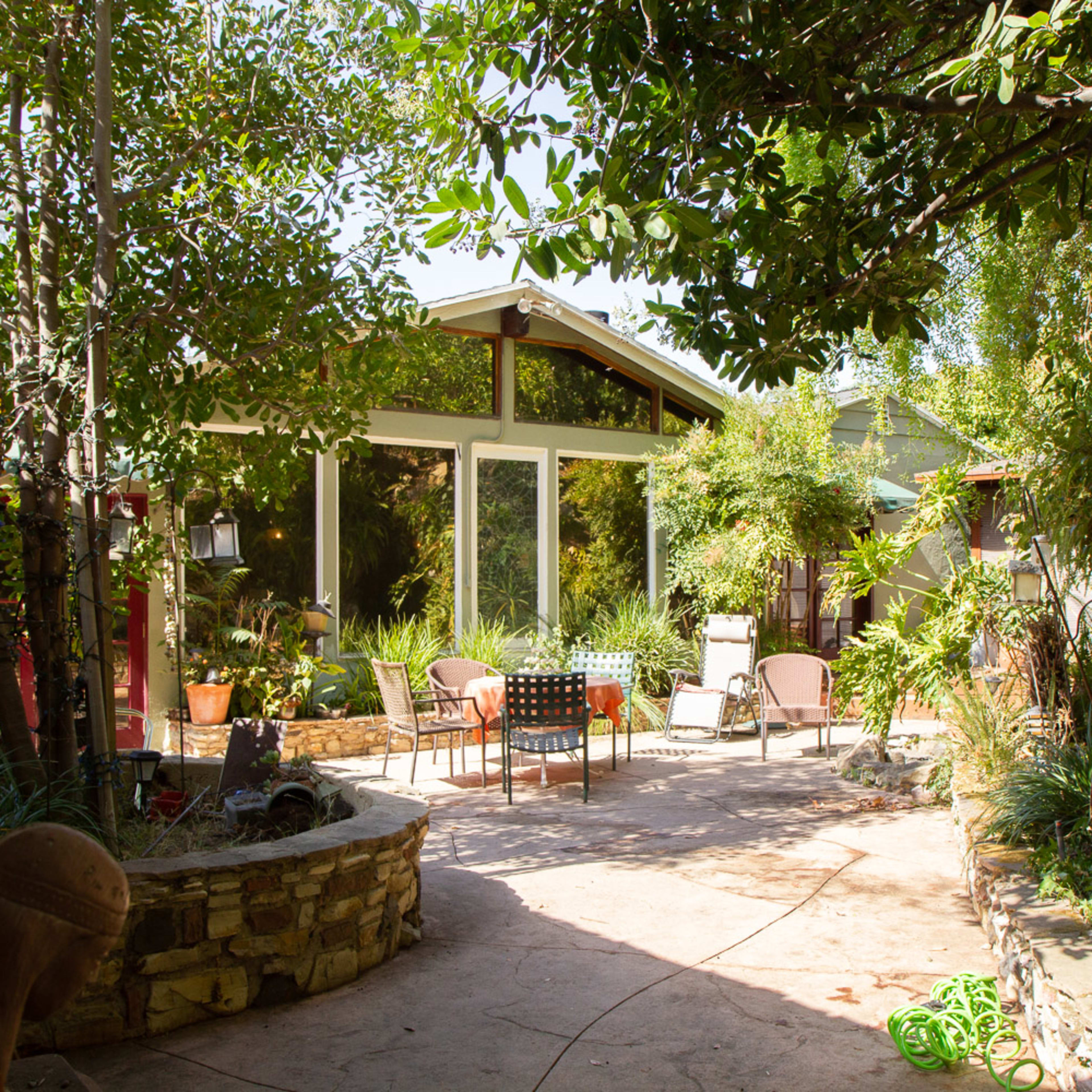 A patio surrounded by greenery features a seating area with chairs and a table, leading to a house with large windows.