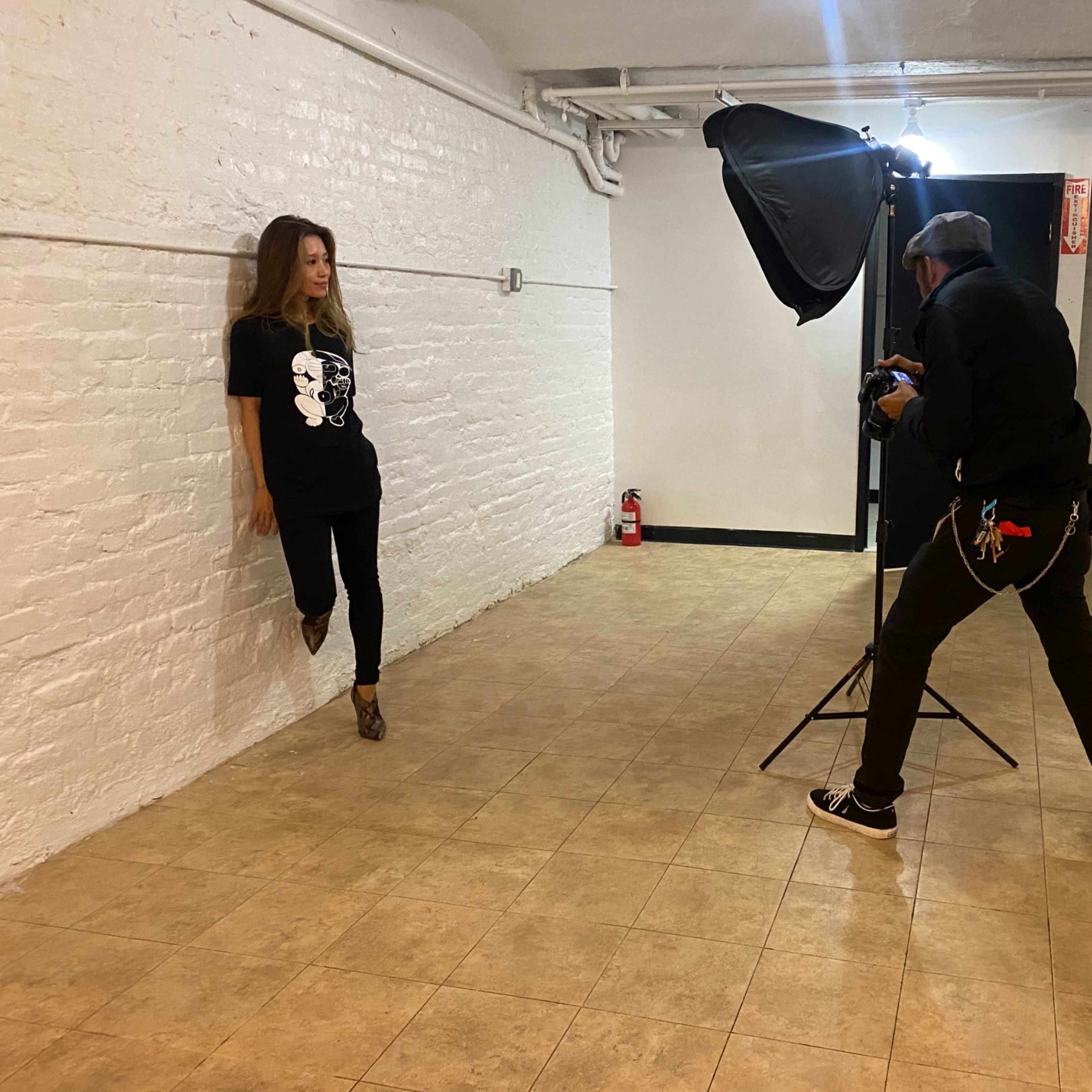 A photographer captures a model posing against a white brick wall in a sparse, well-lit studio space.