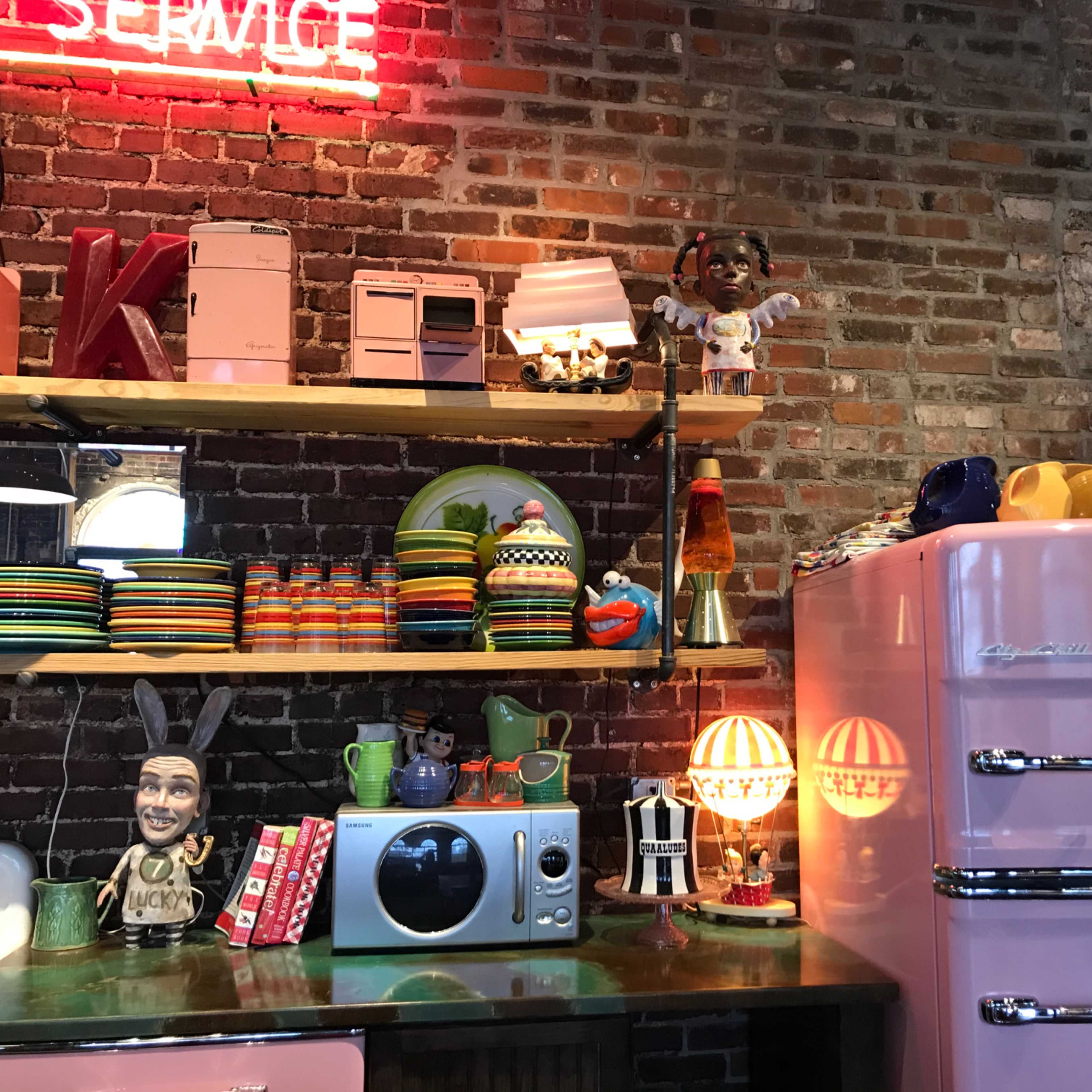 A vintage kitchen setup with a pink refrigerator, shelves filled with colorful dishes and decorative items, and a brick wall in the background.