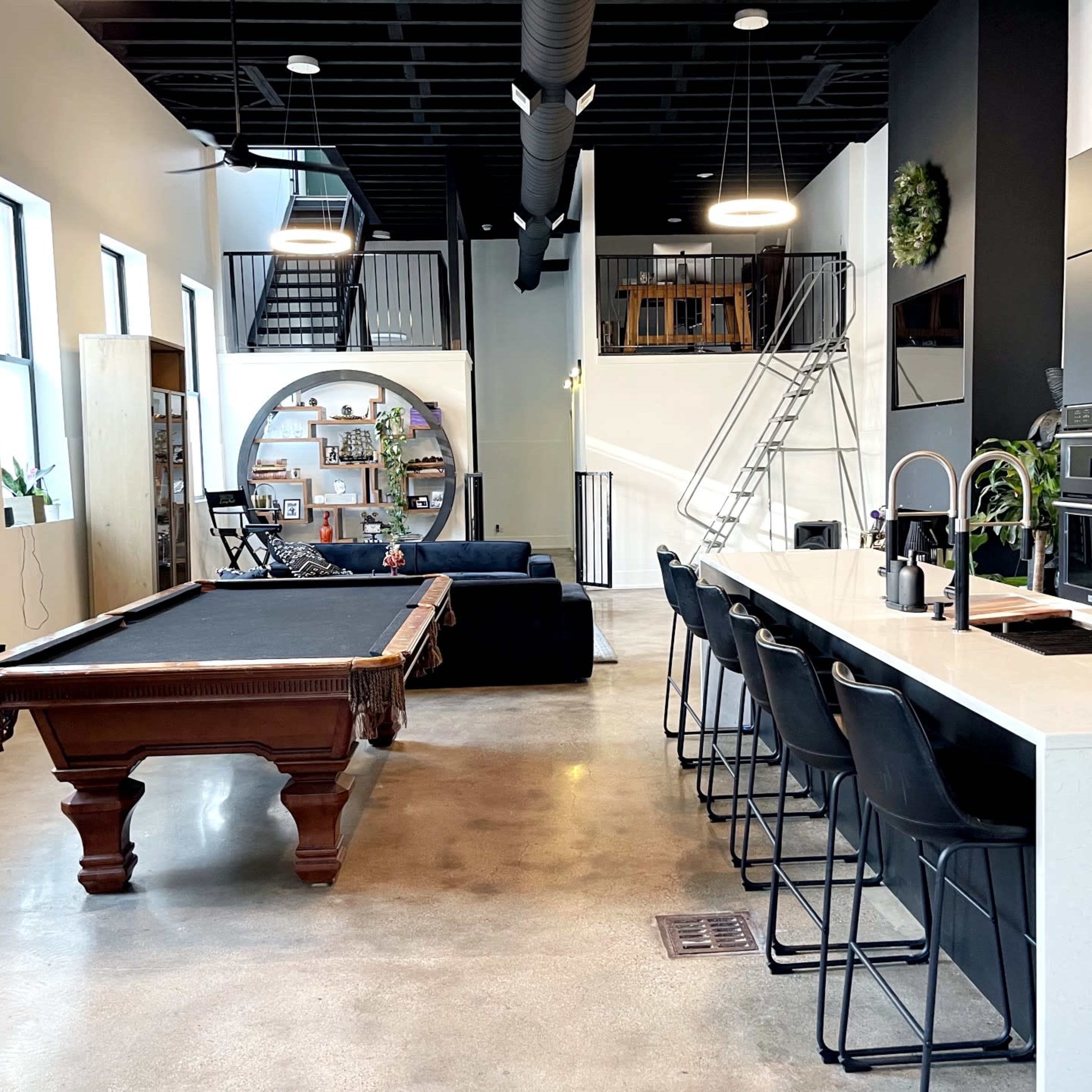 A modern open-concept living space featuring a pool table, a kitchen island with seating, and a staircase leading to an upper level.