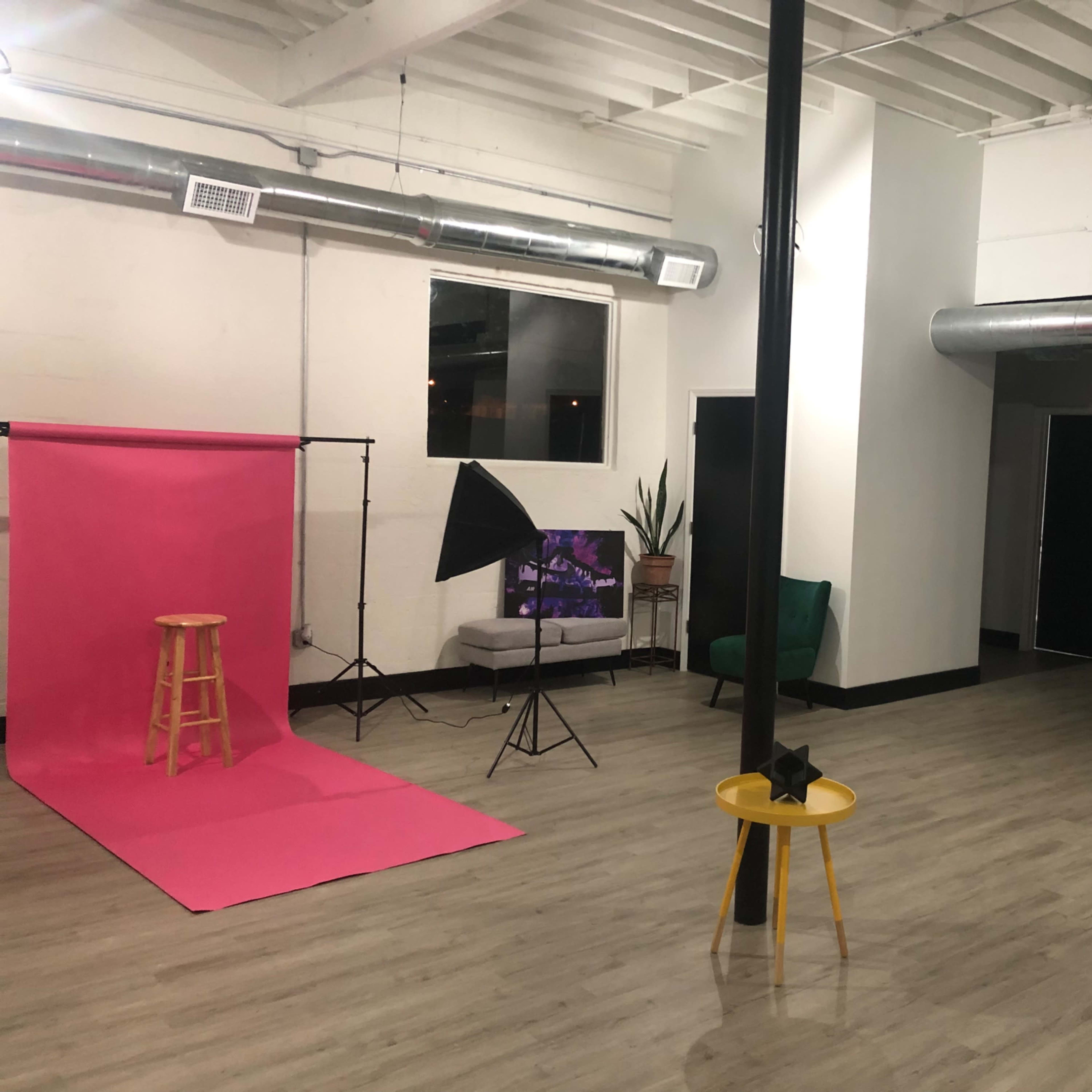 A spacious studio with a pink backdrop, a stool, a photography light, and a green armchair, along with a small black and white shelf in the corner.