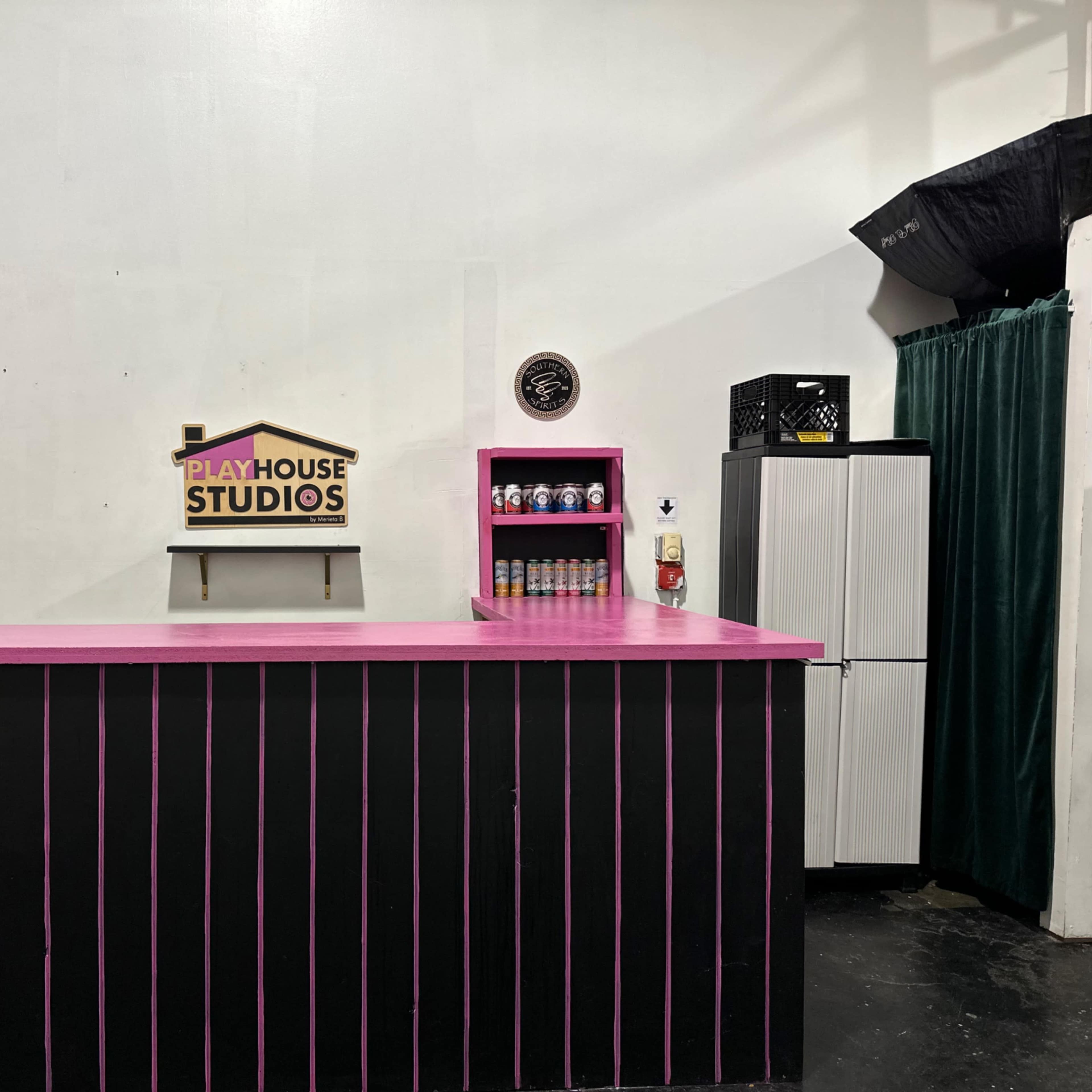 A studio interior with a pink and black counter, a sign that reads "PLAYHOUSE STUDIOS," a refrigerator, and a black curtain in the background.