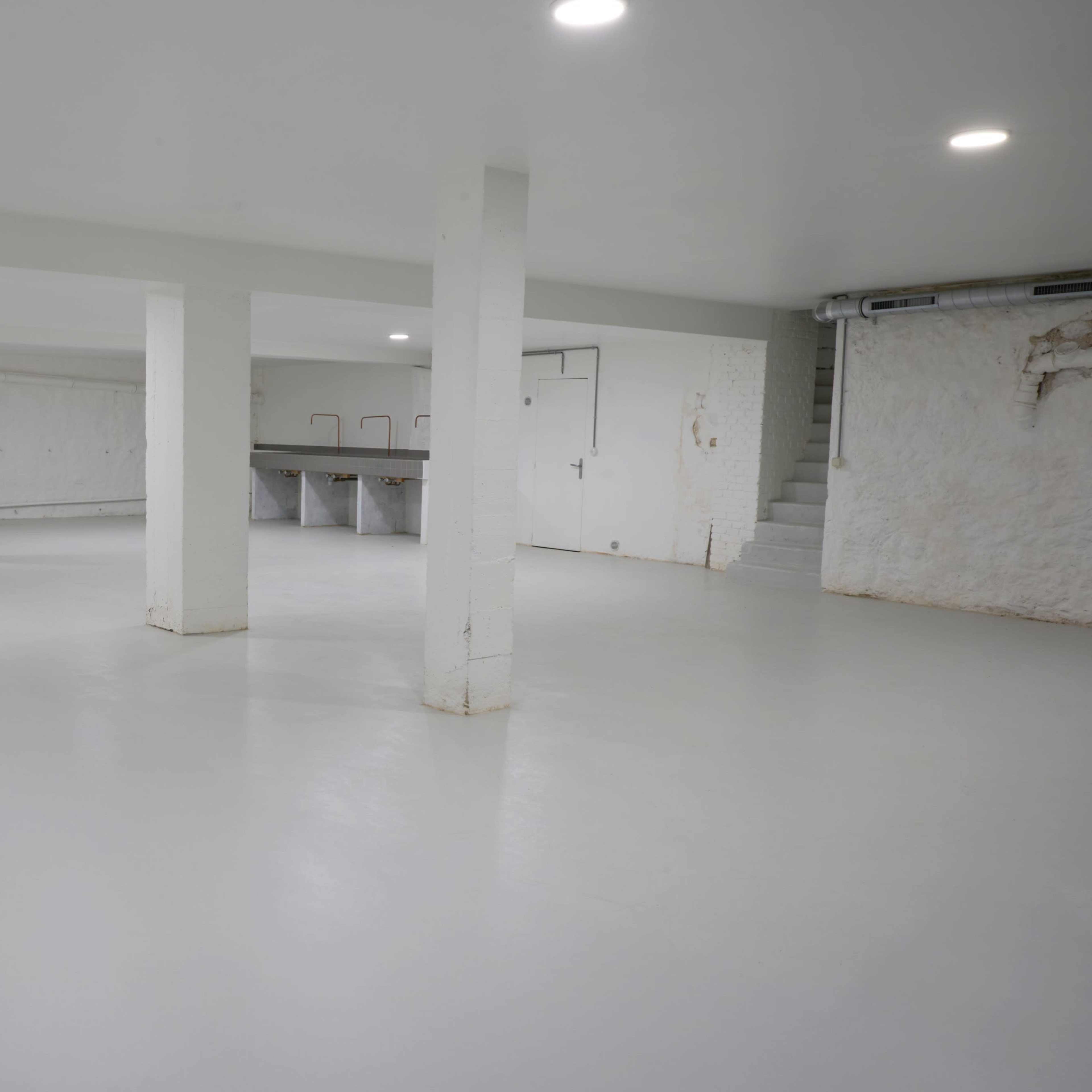 A spacious, empty basement with white walls and a smooth white floor, featuring several support columns and a staircase in one corner.