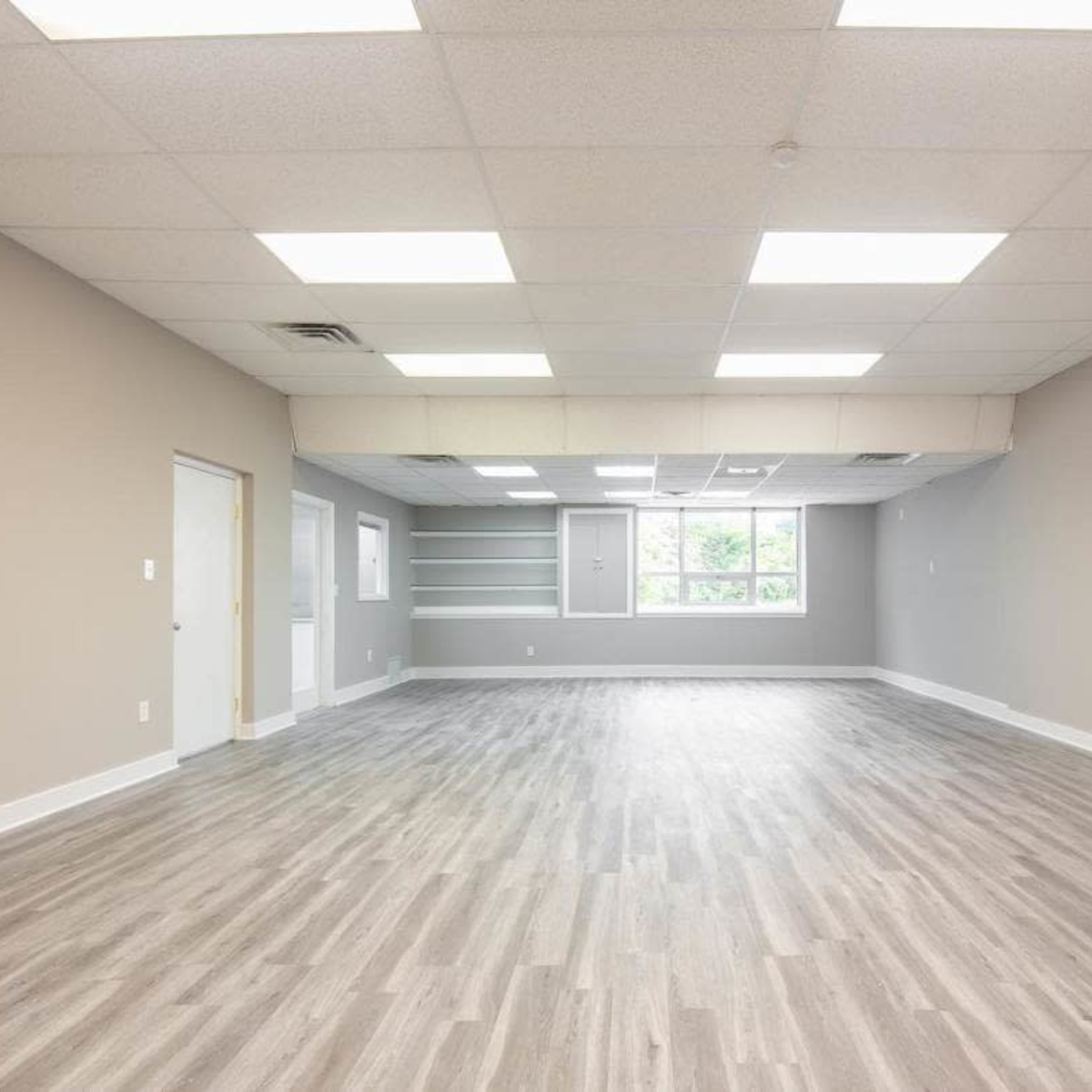 A spacious, empty office interior with gray walls, a wood-like floor, and multiple overhead lights.
