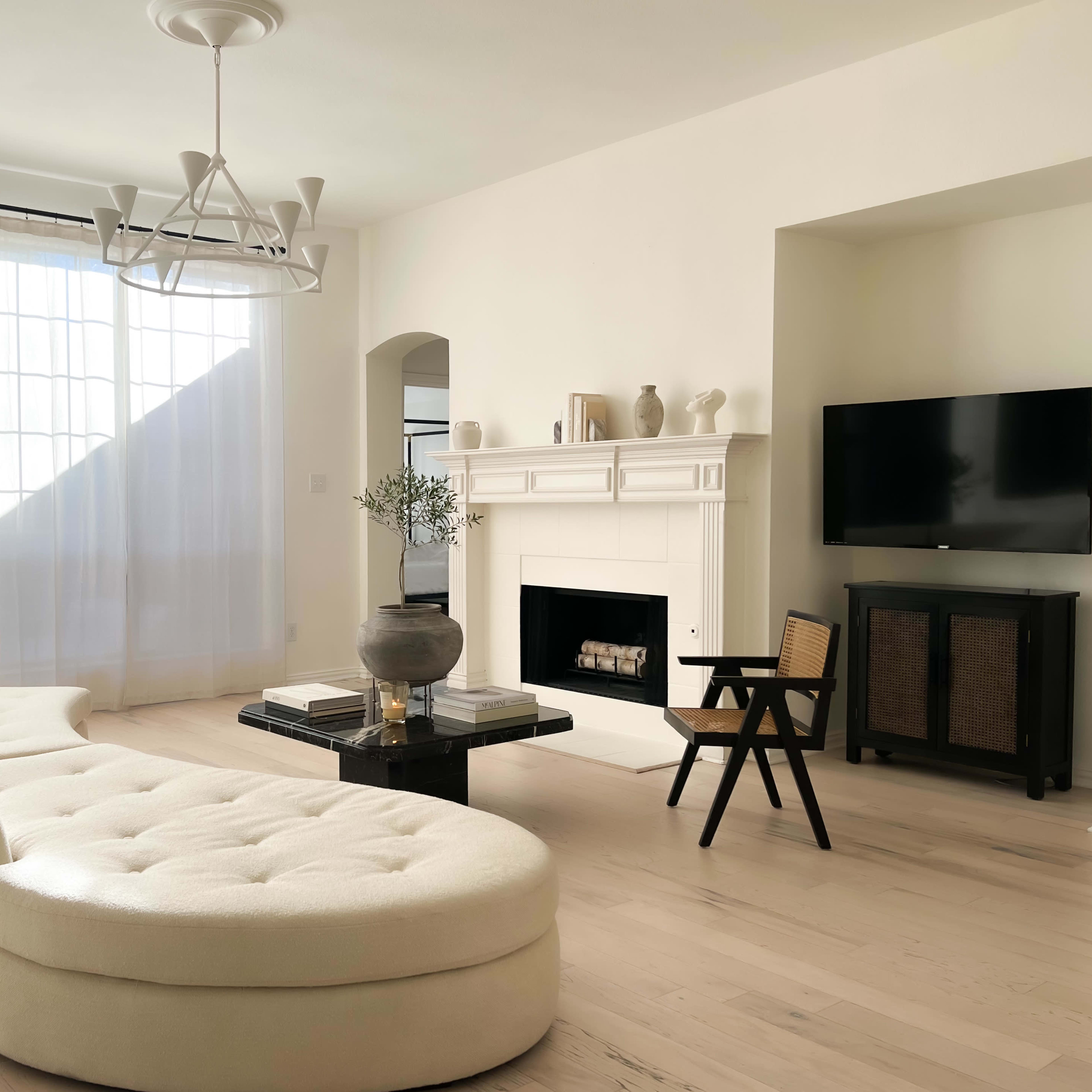 A modern living room features a white curved sofa, a black coffee table with a decorative vase, a fireplace, and a television against a light-colored wall.