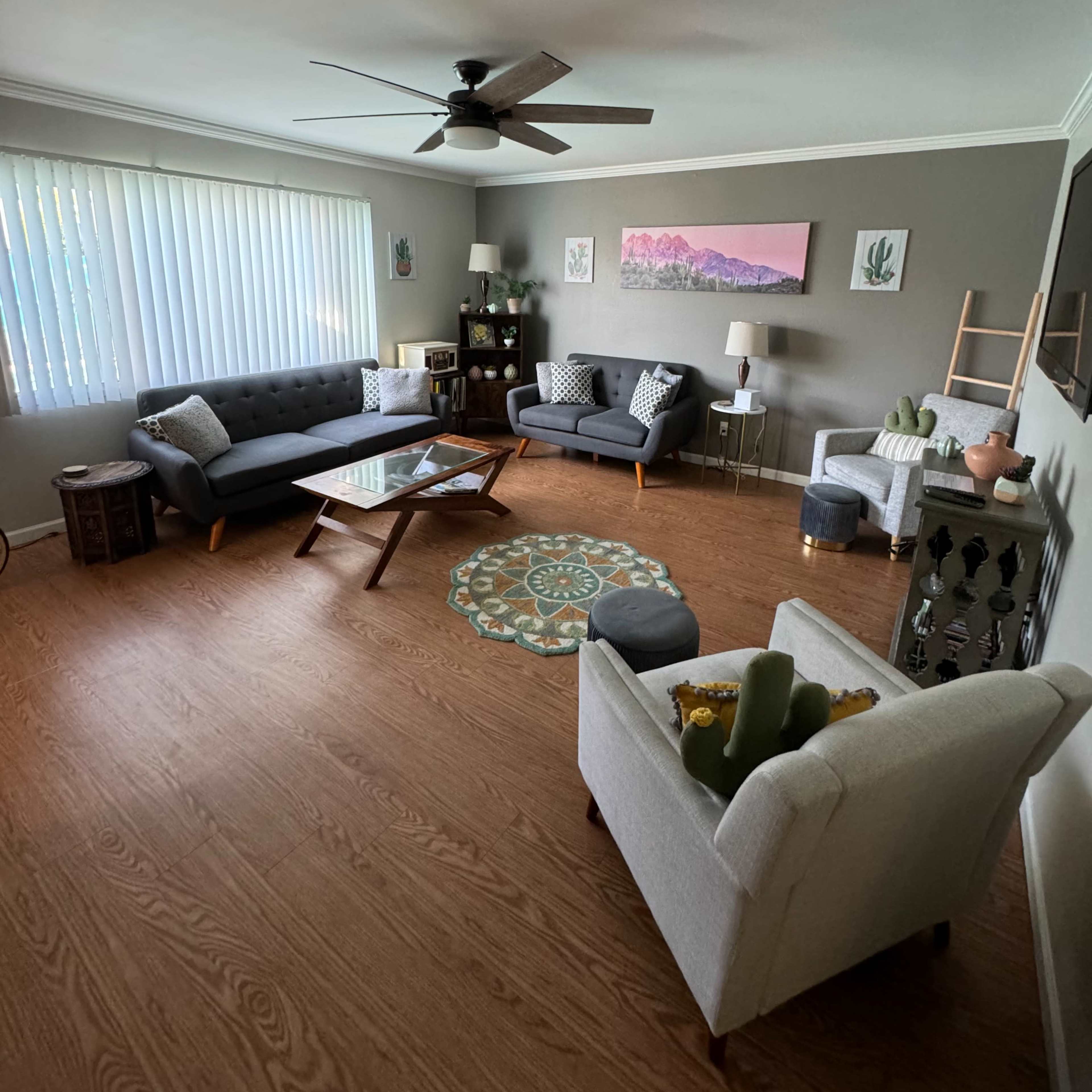 A living room features two gray sofas, a glass coffee table, a ceiling fan, and decorative items, with a large window allowing natural light.