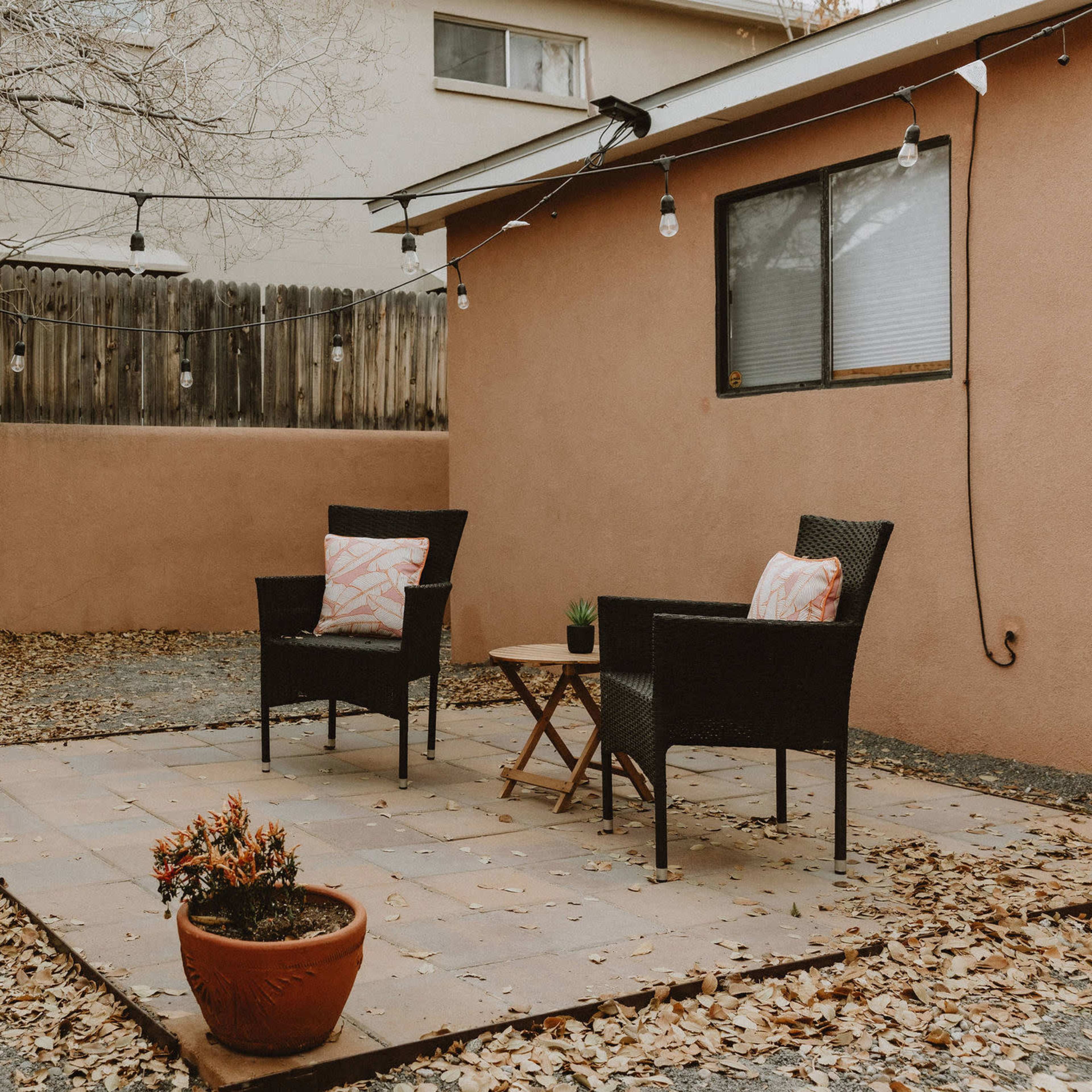 A patio area features two black chairs with cushions, a small wooden table, and a potted plant, surrounded by dry leaves and a tree in the background.