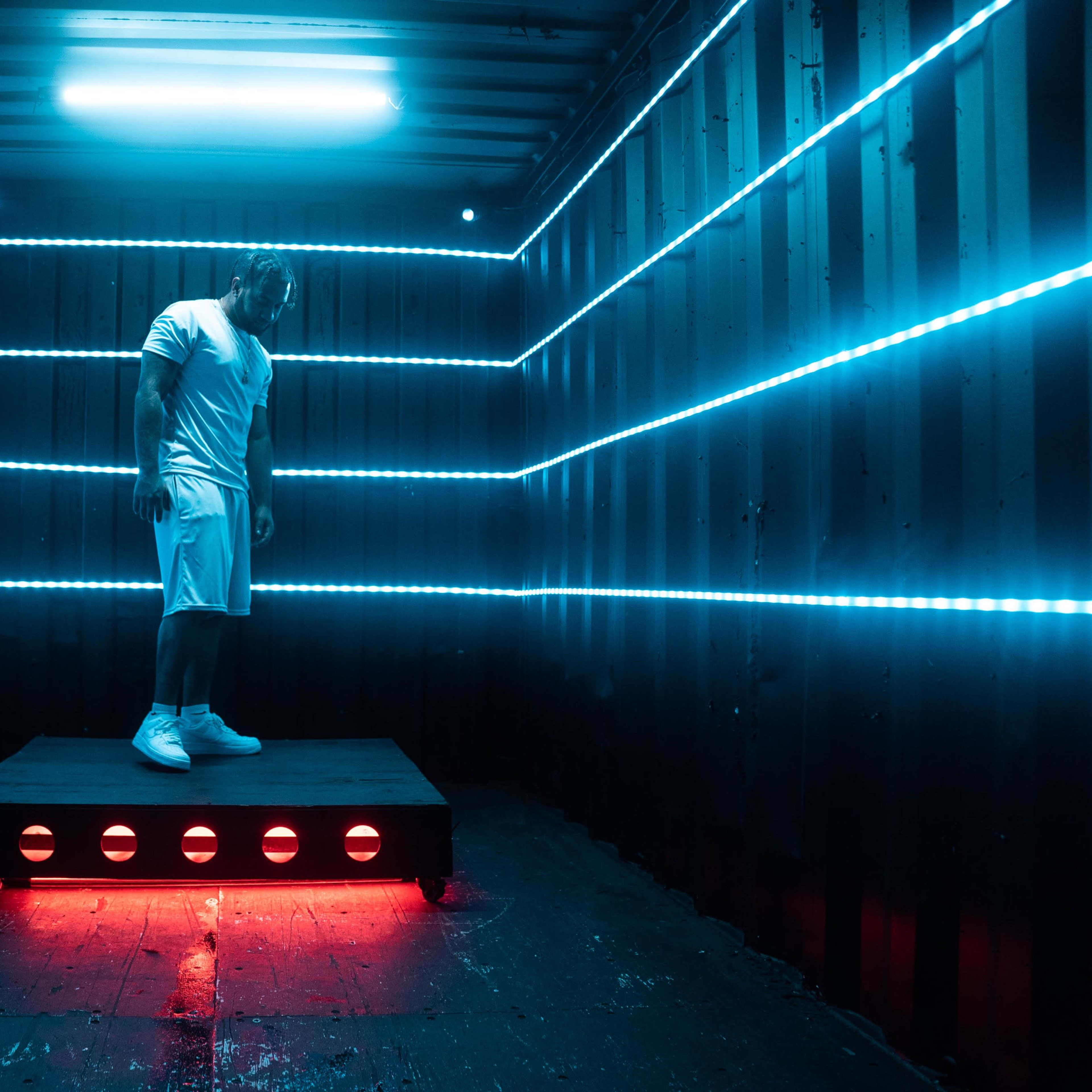A figure stands on a raised platform inside a dimly lit shipping container, illuminated by blue and red LED lights.