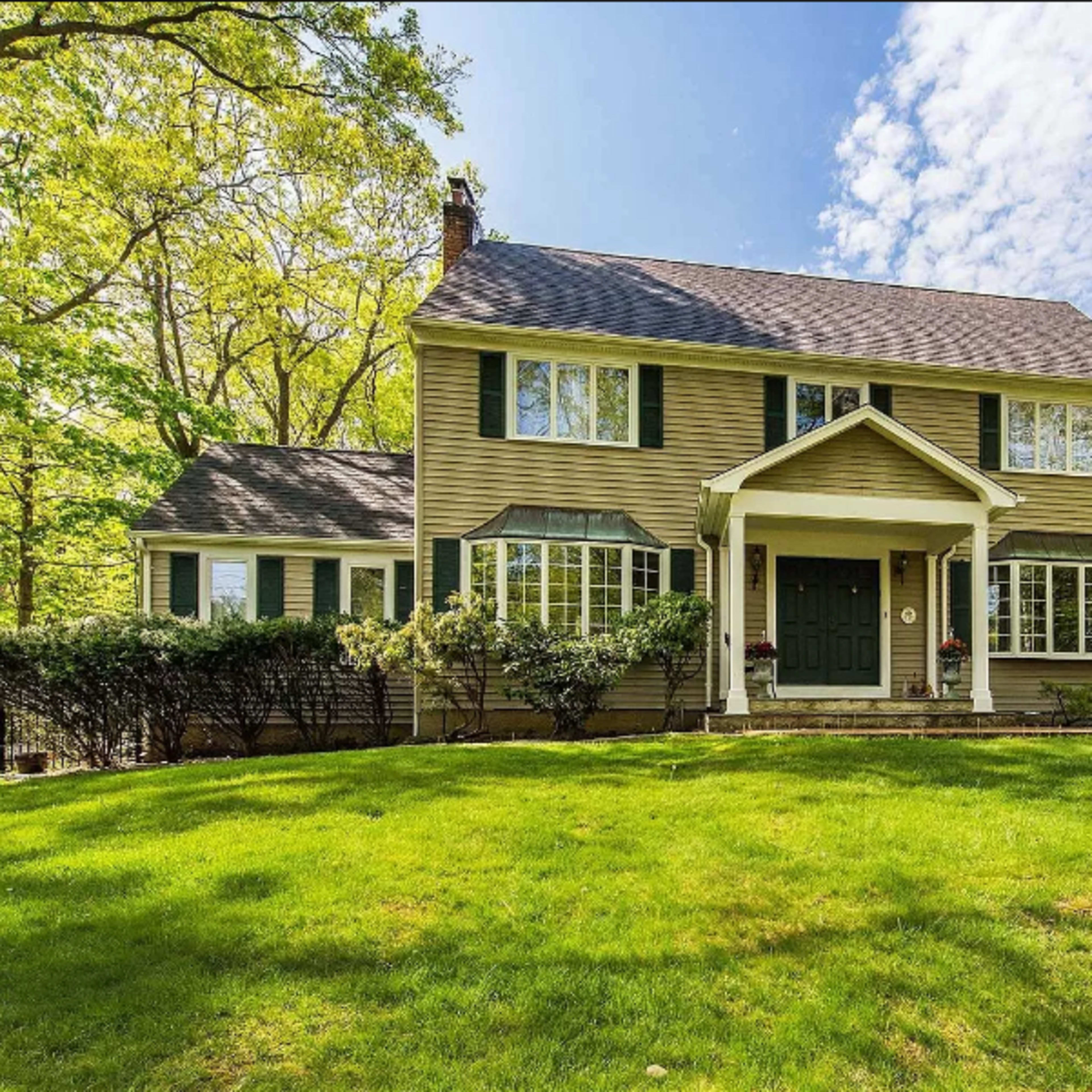 A two-story house with green shutters and a large front lawn is surrounded by trees and features a covered entrance.