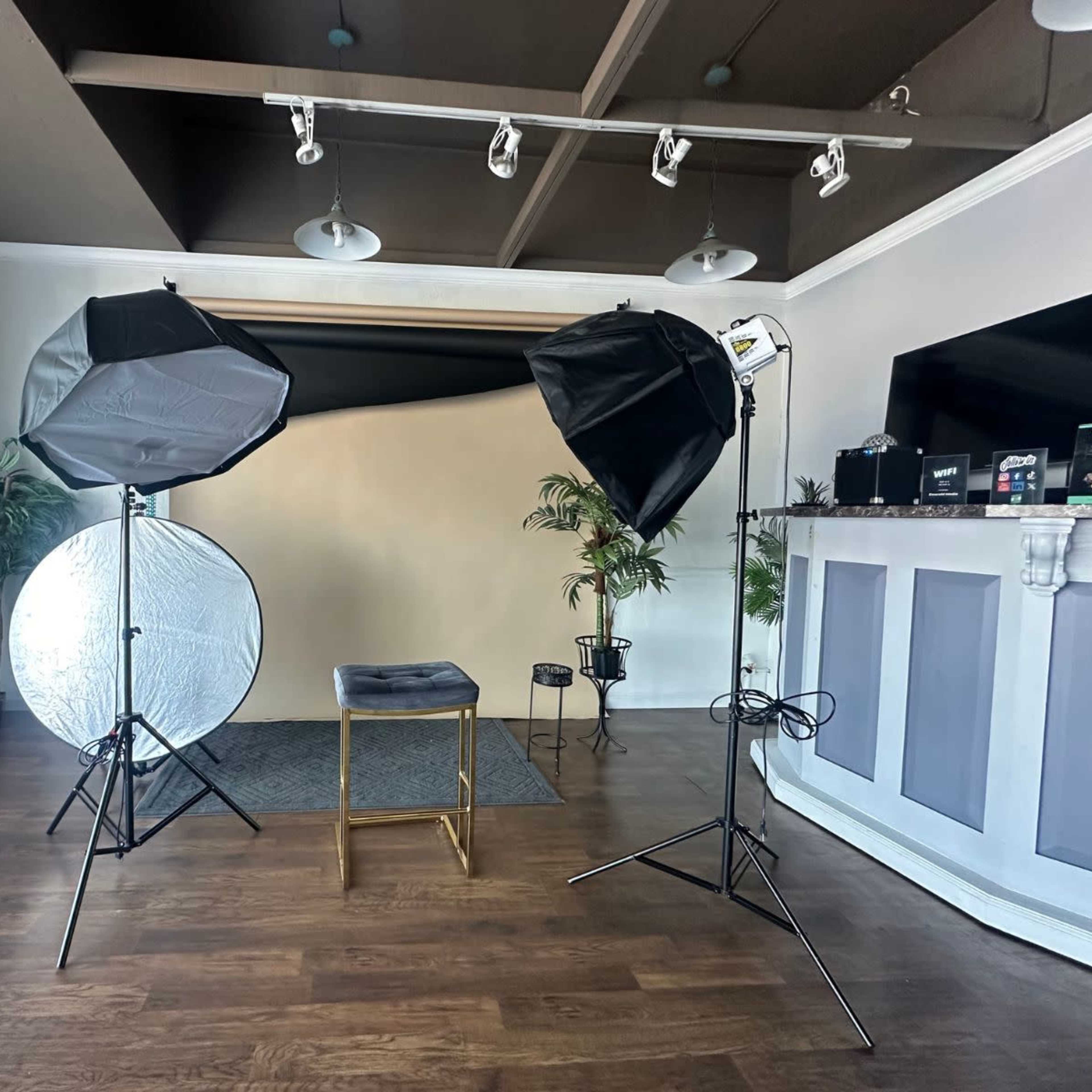 A photography studio setup with two softboxes, a reflective umbrella, a stool, and a backdrop on a wooden floor.