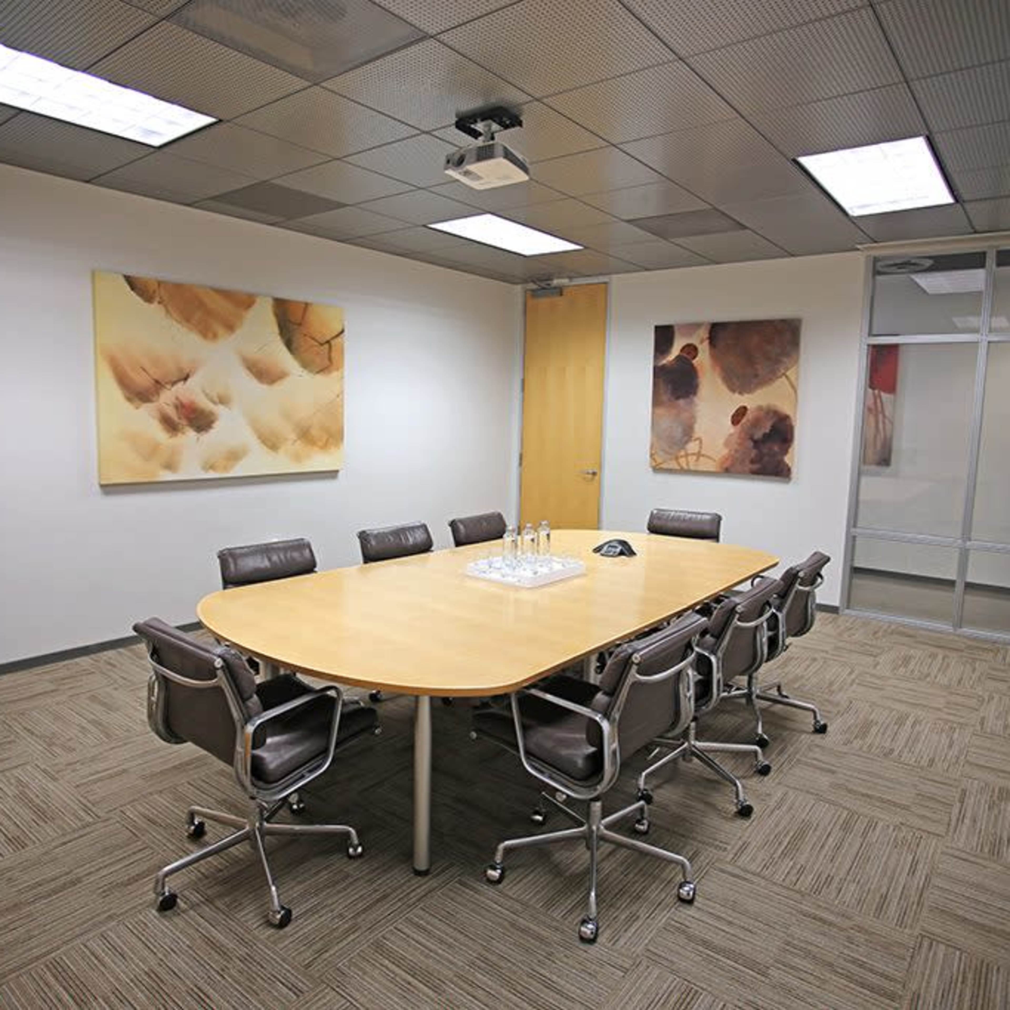 A conference room features a large oval table surrounded by six chairs, with two abstract paintings on the walls and a projector mounted on the ceiling.