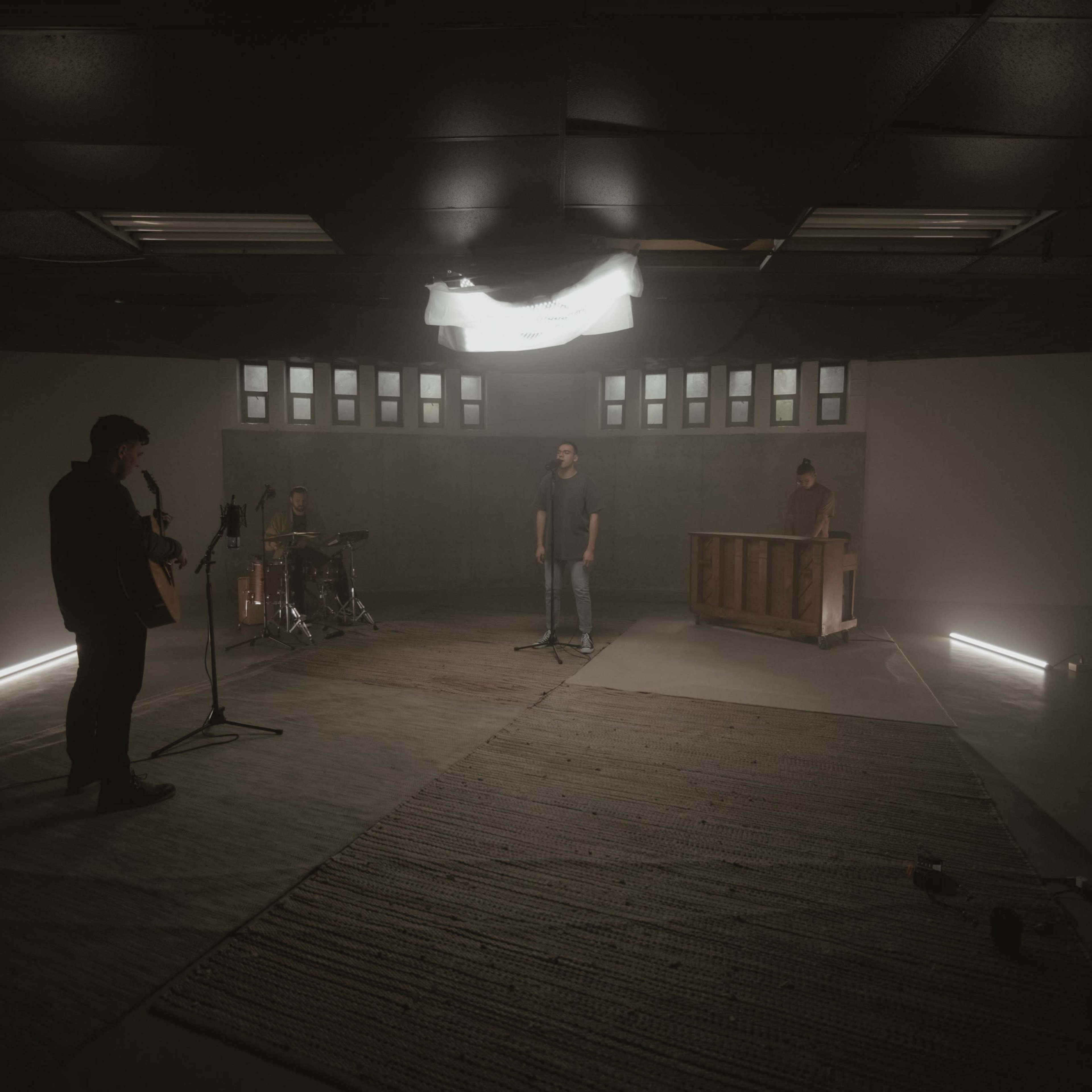 A band performs in a dimly lit rehearsal space featuring instruments like a guitar, drums, and a piano.