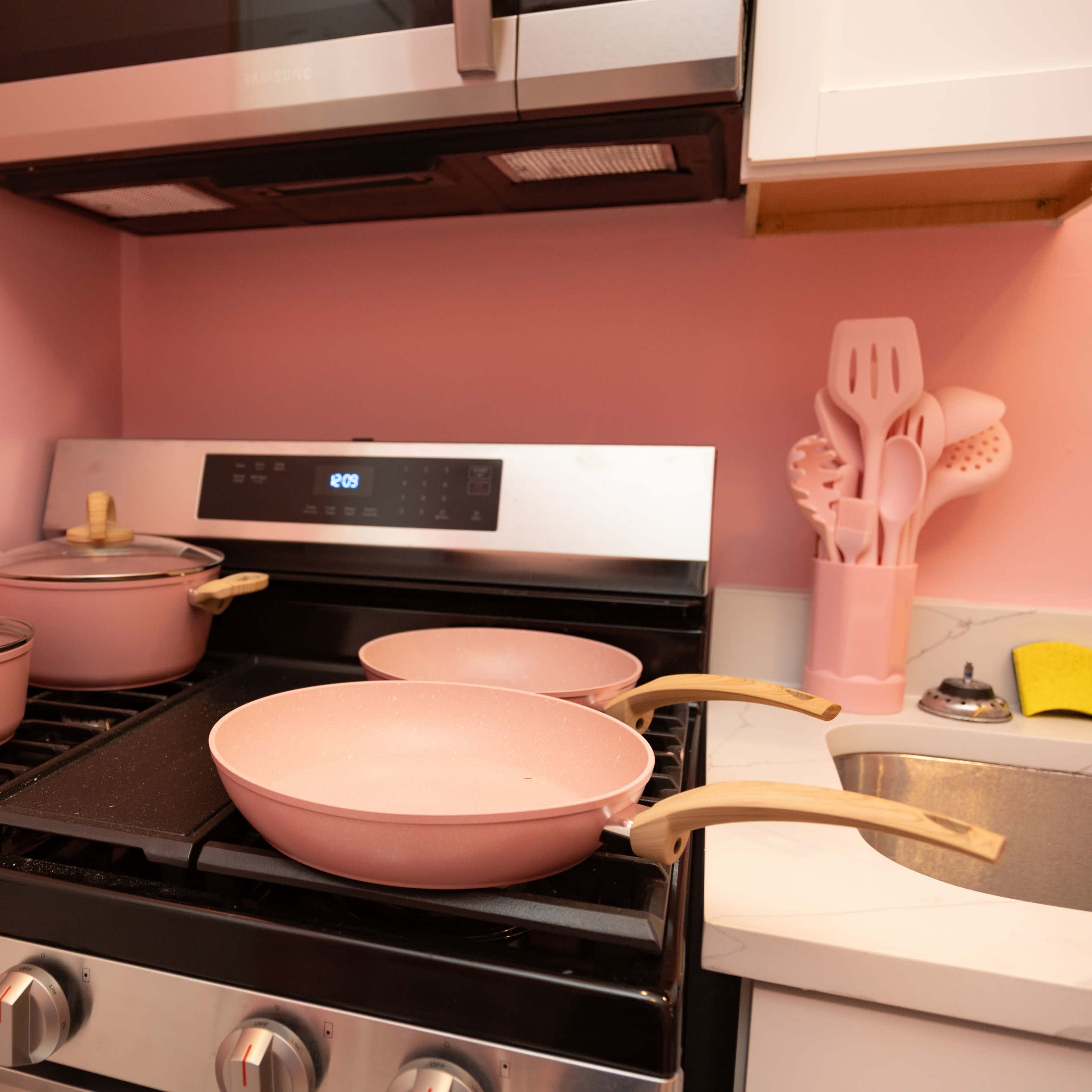 A pink-themed kitchen features a stove with two frying pans and two pots, along with cooking utensils in a matching color.