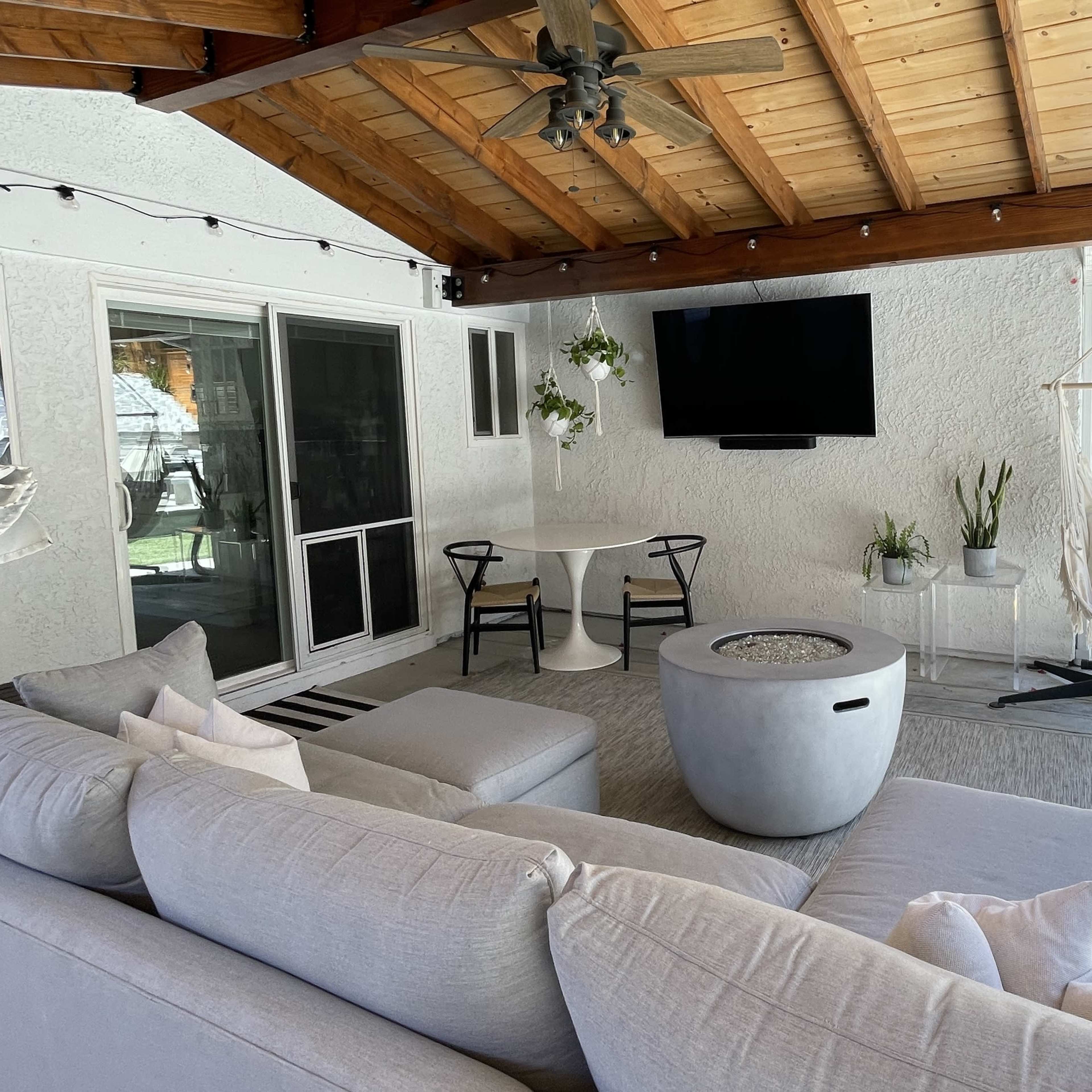 A covered outdoor living area featuring a gray sectional sofa, a round table, a television mounted on the wall, and a hanging chair.