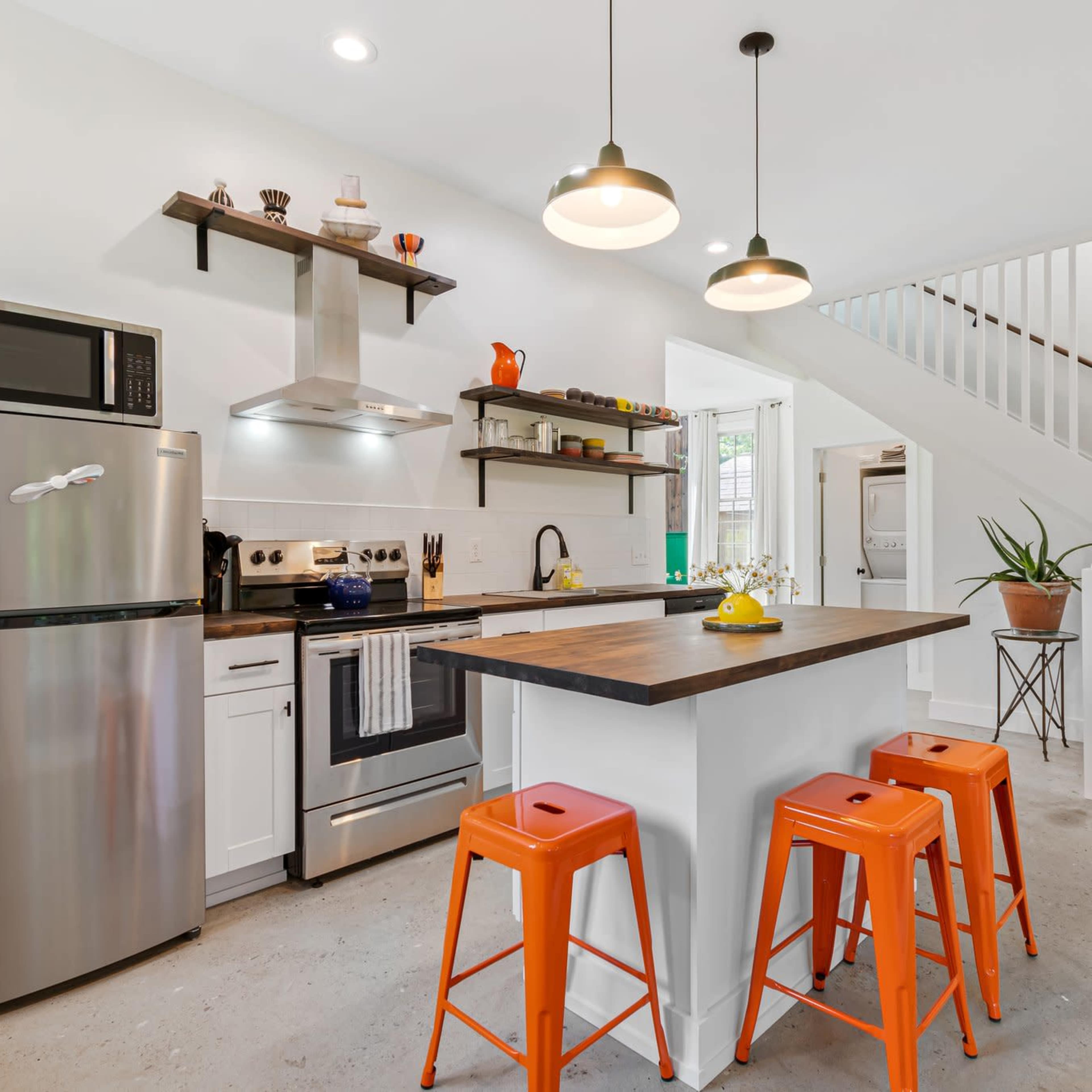 A modern kitchen with stainless steel appliances, a central island with orange stools, and open shelving displaying decorative items.