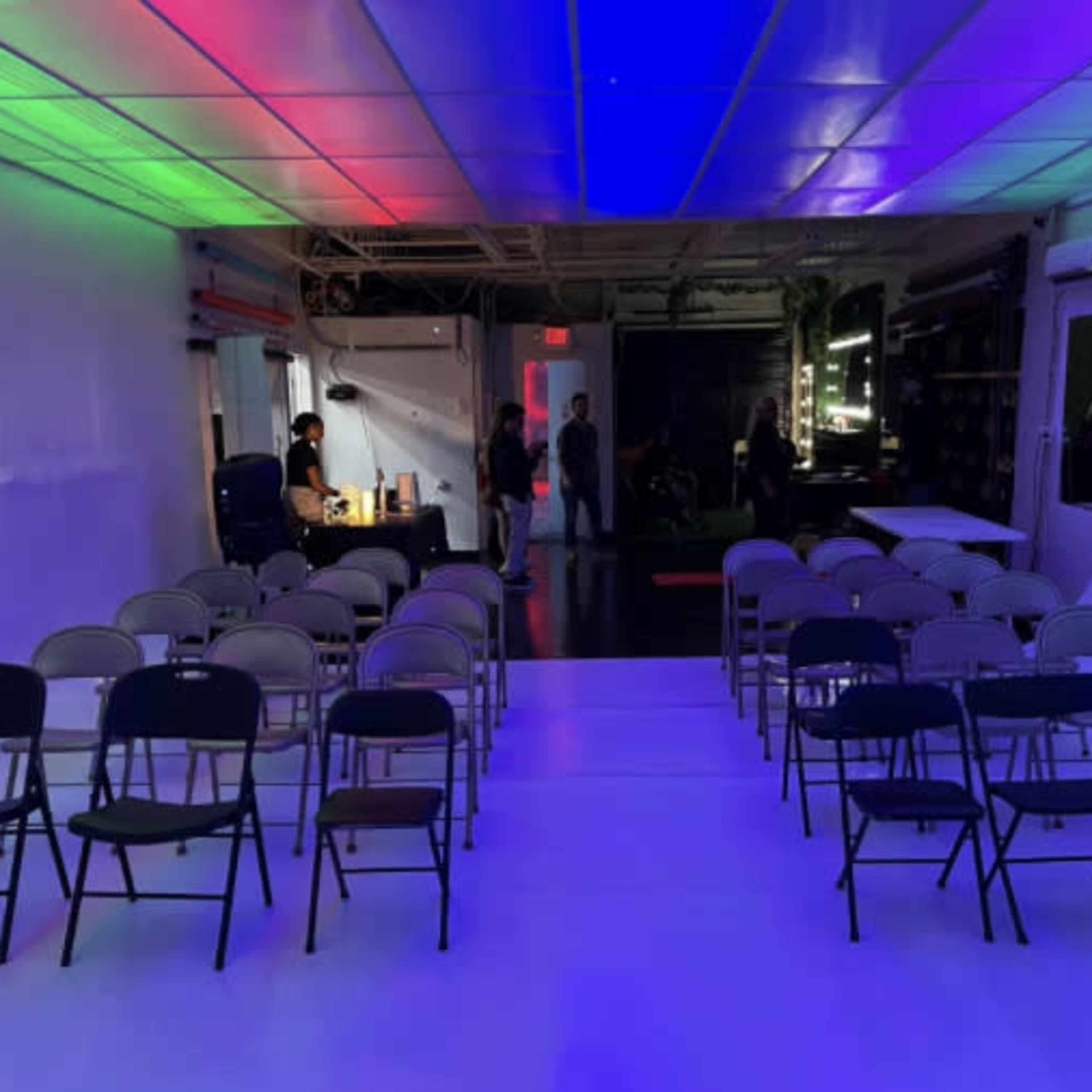 A spacious room with rows of folding chairs set up for an event, illuminated by colorful lights on the ceiling.