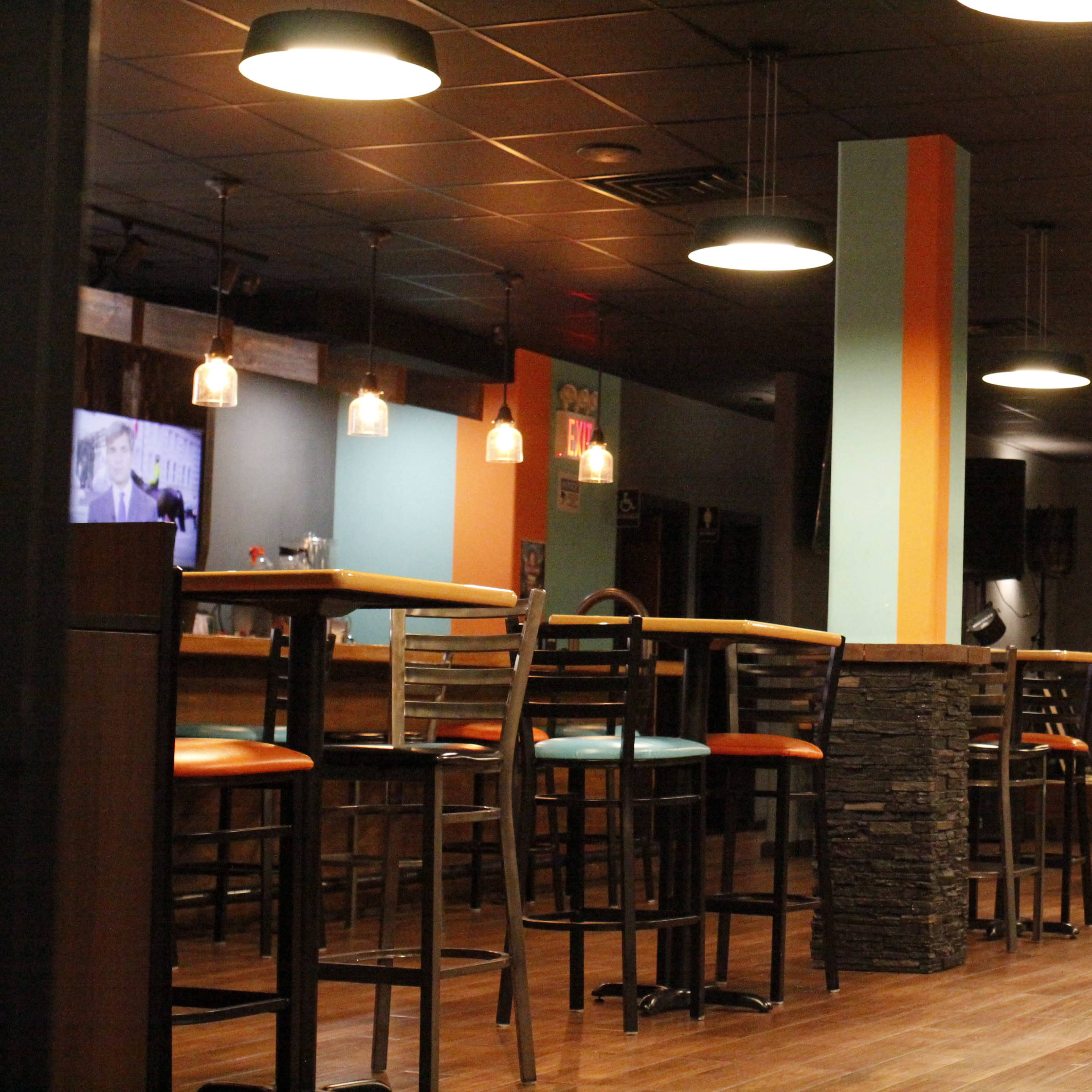 The interior of a bar with high stools and tables, illuminated by pendant lights.