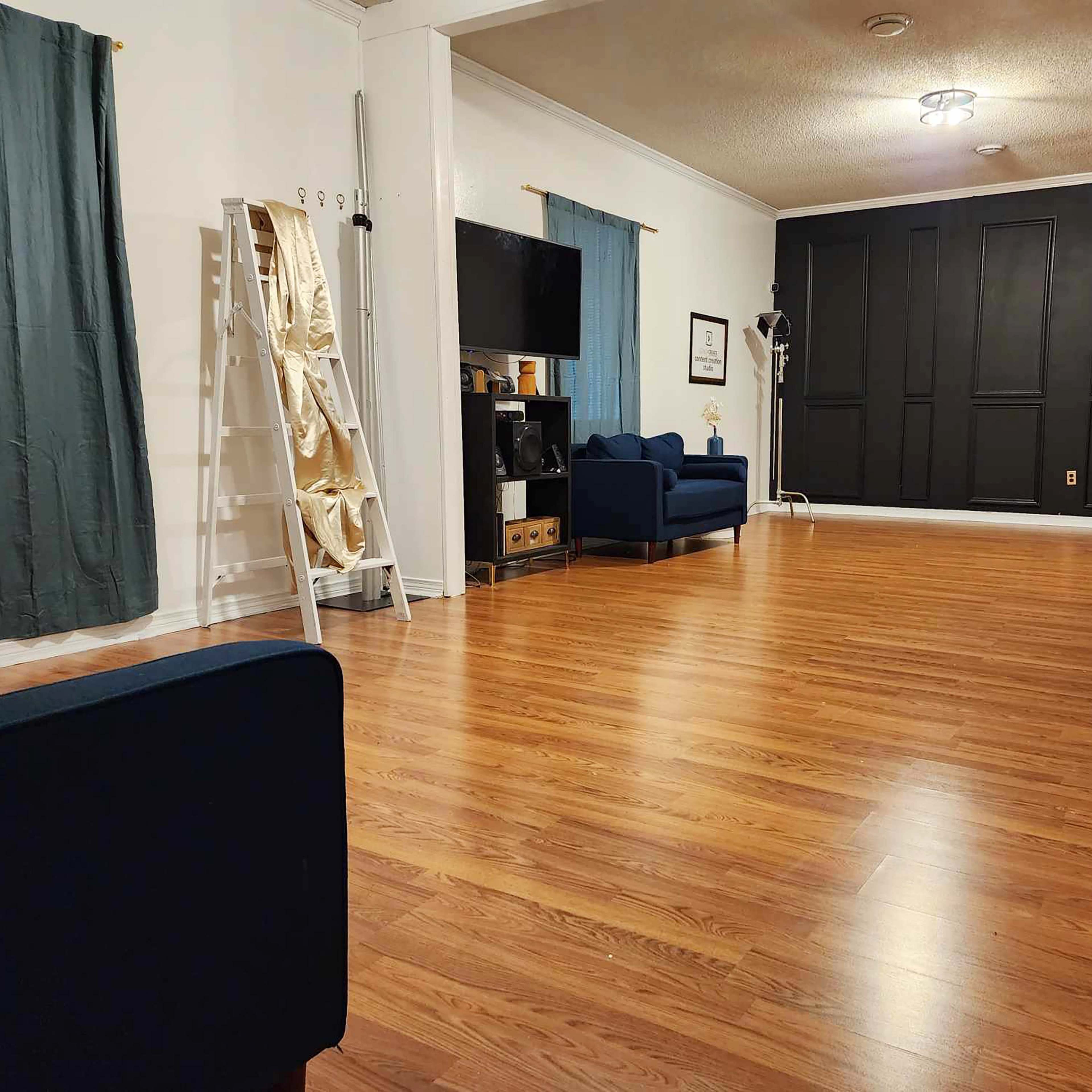 The room features wooden flooring, two blue couches, a black accent wall, and tall windows covered with curtains.