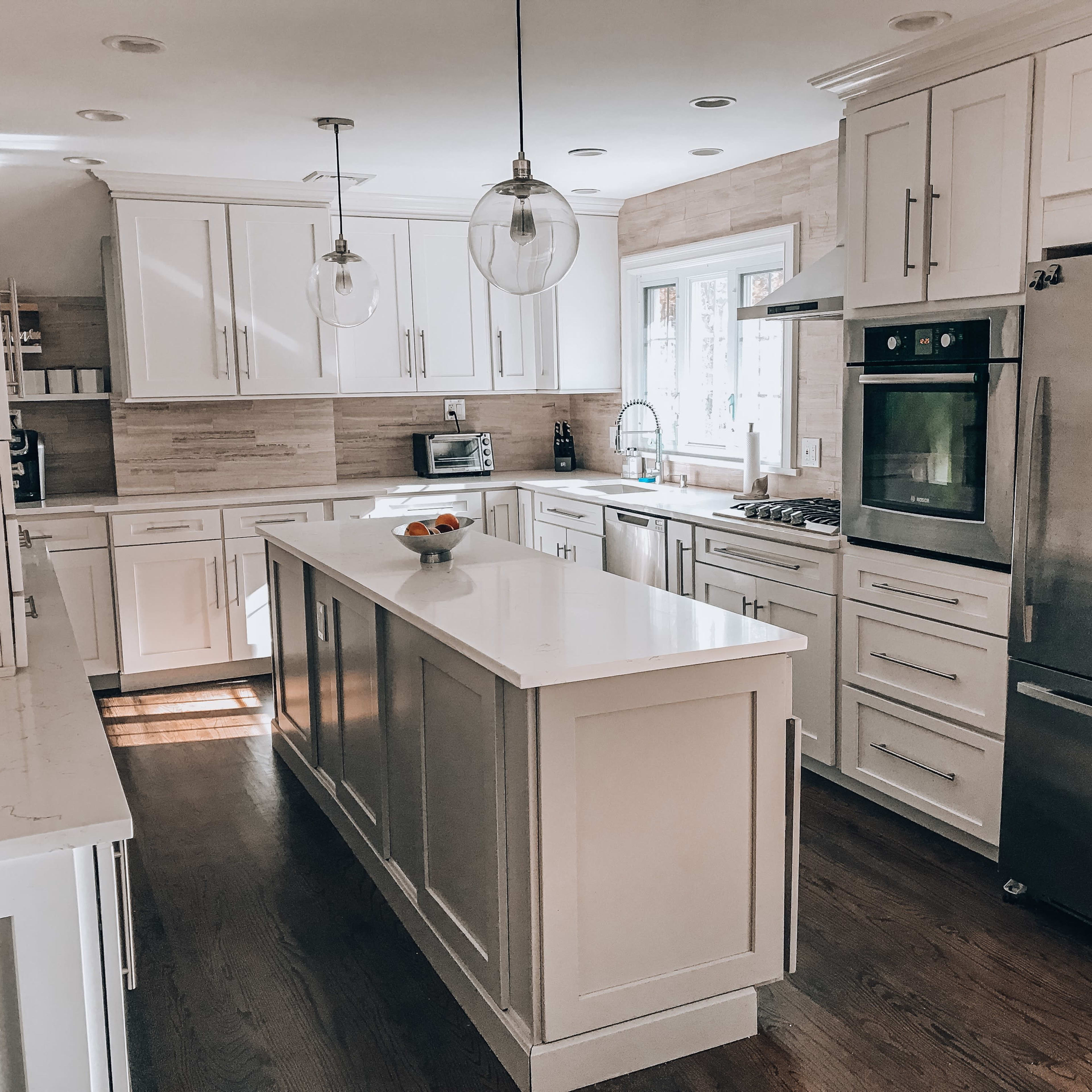 A modern kitchen with white cabinets, a central island, stainless steel appliances, and a large window providing natural light.