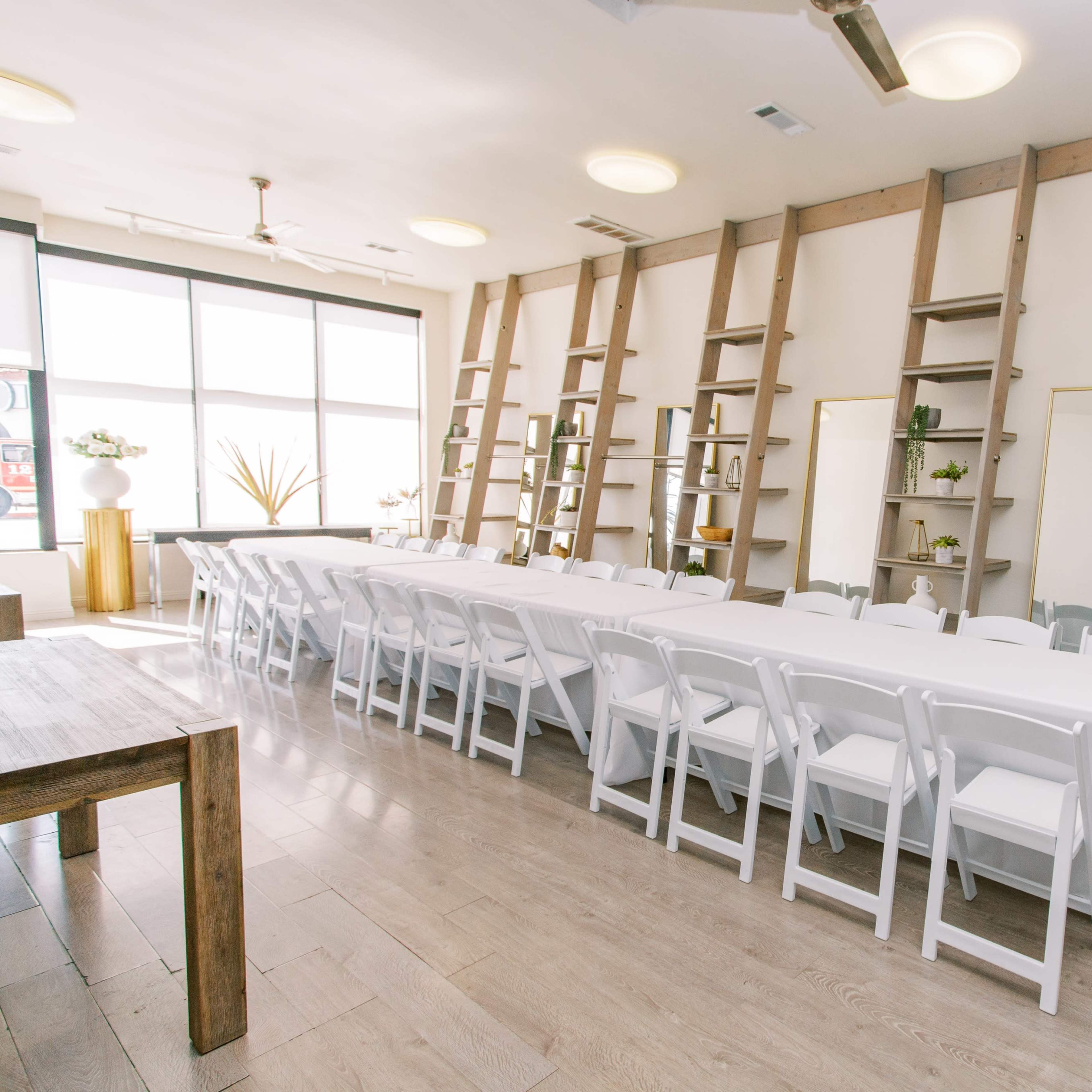 A long, white table is set with chairs in a well-lit room featuring wooden shelves on the walls and plants arranged decoratively.
