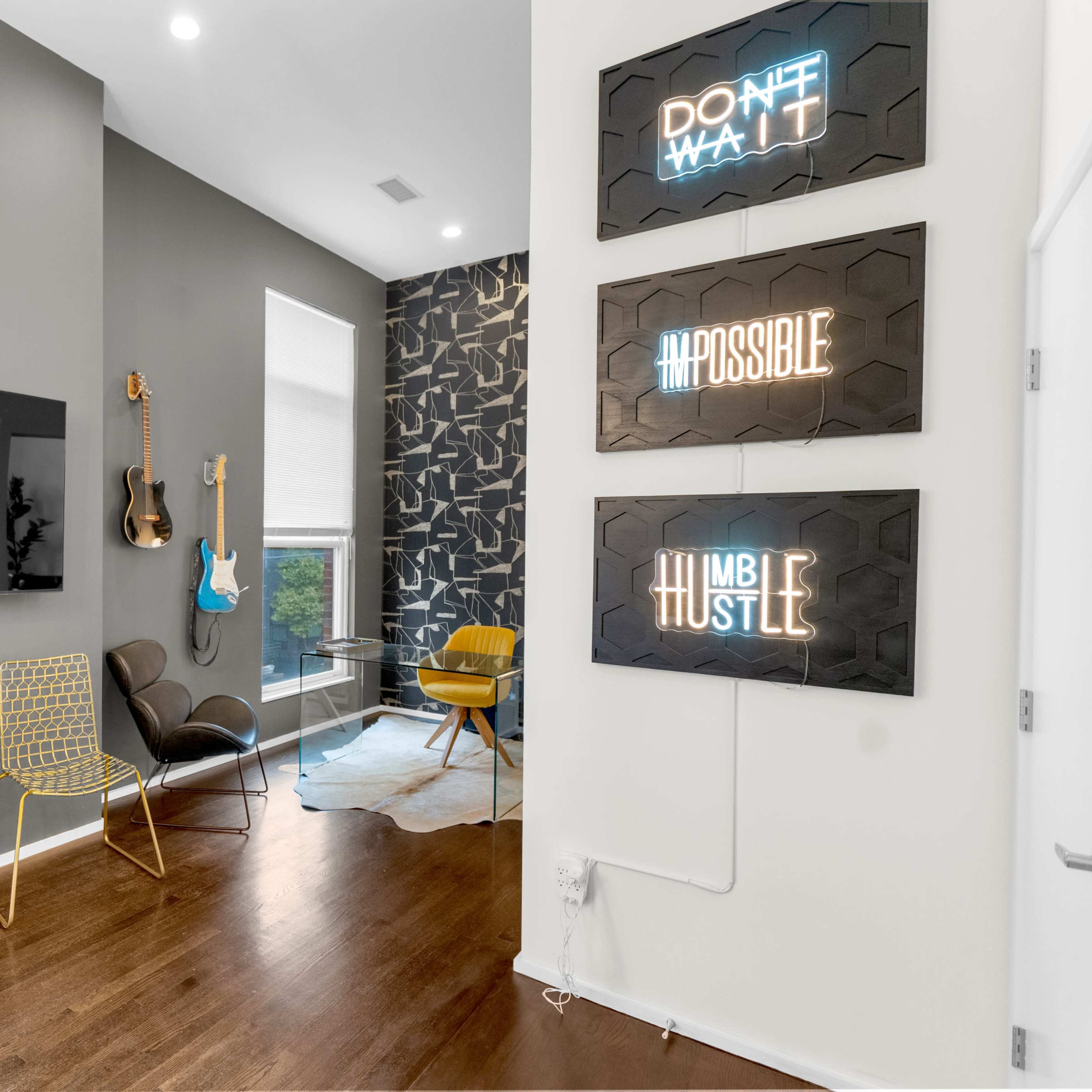 A modern room with a television mounted on the wall, two styled chairs, a guitar on display, and inspirational neon signs framed on the wall.